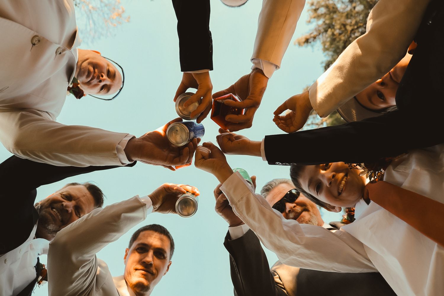 Group of men toasting with cans in a circle, viewed from below against a blue sky at a celebration.