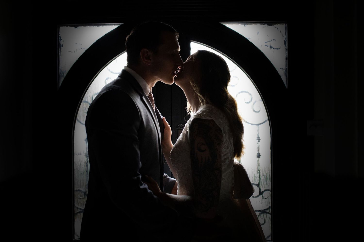 Romantic silhouette of a couple kissing in front of an ornate arched window on their wedding day.