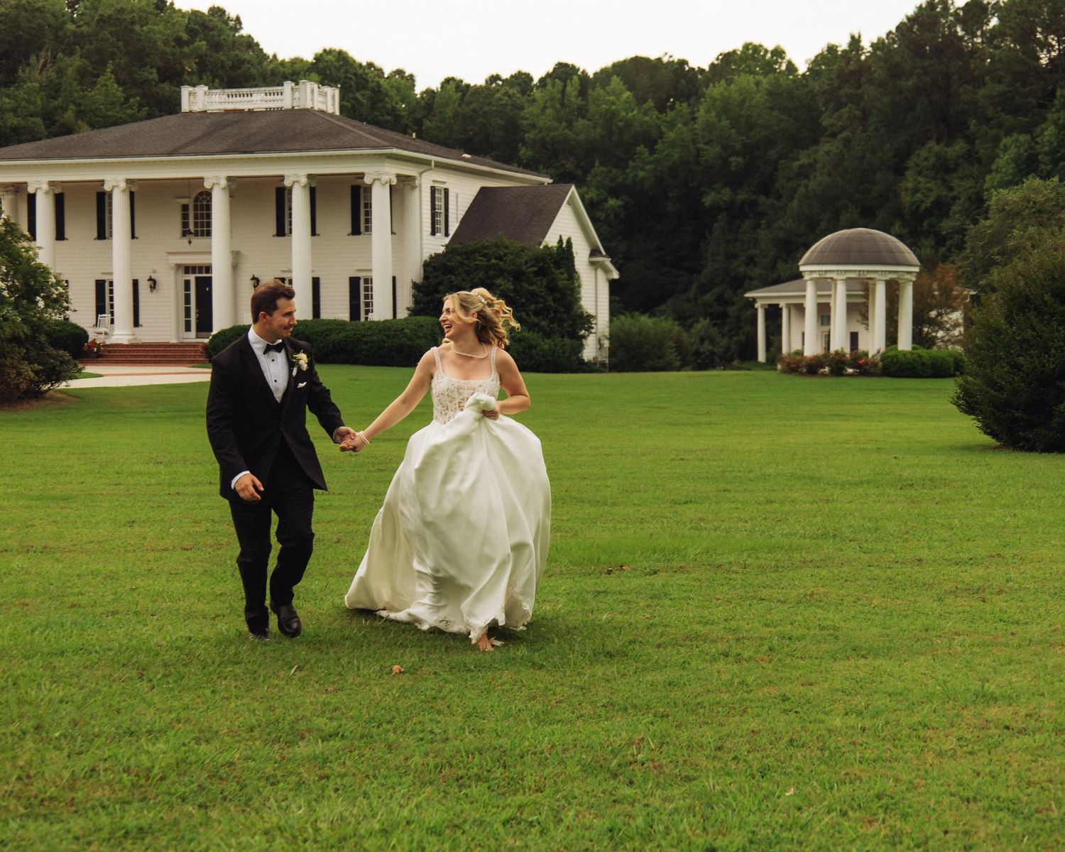 Bride and groom holding hands running across green lawn with white plantation-style wedding venue in background.