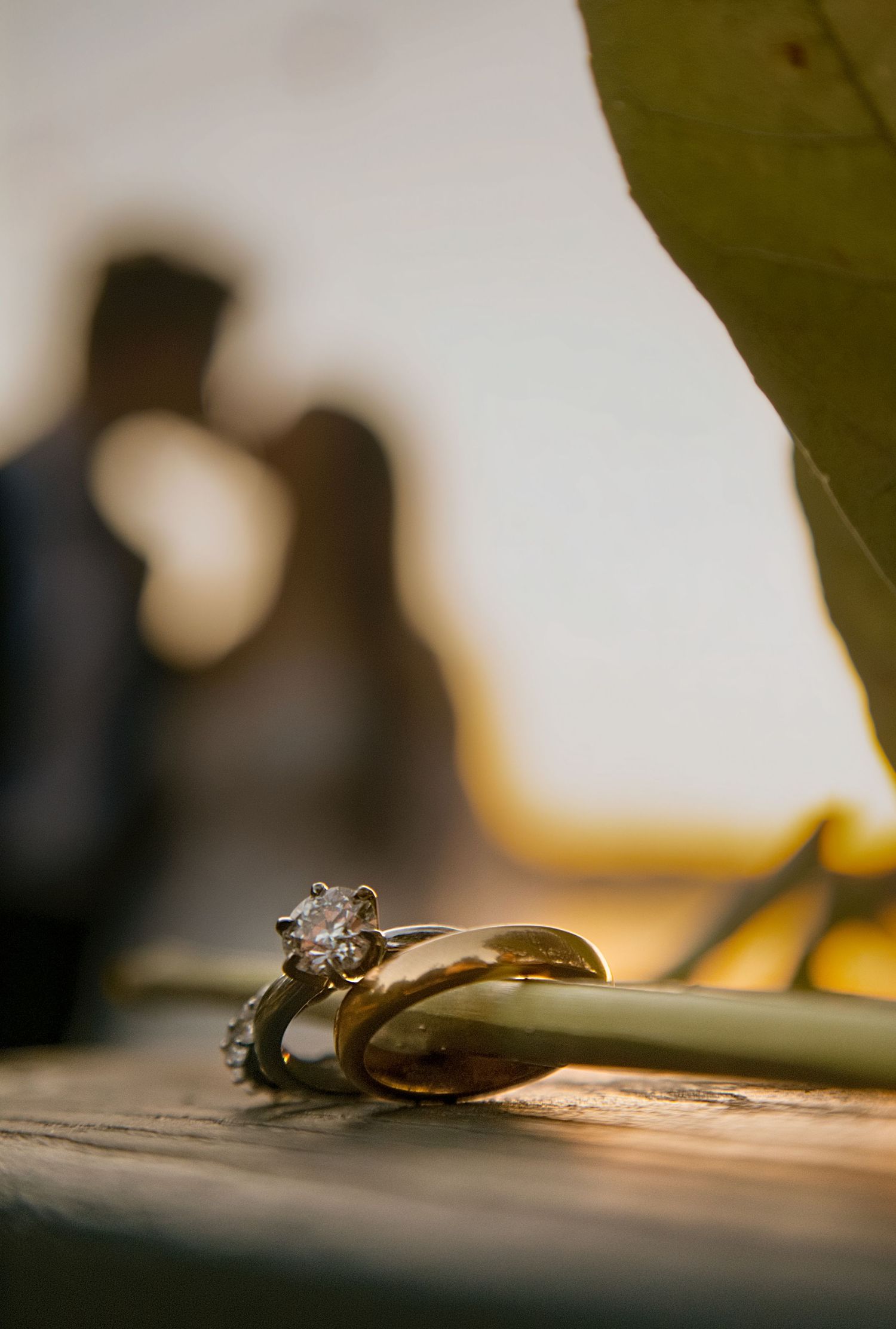 Gold wedding bands and diamond engagement ring rest on a surface with blurred couple silhouette in warm sunset background.