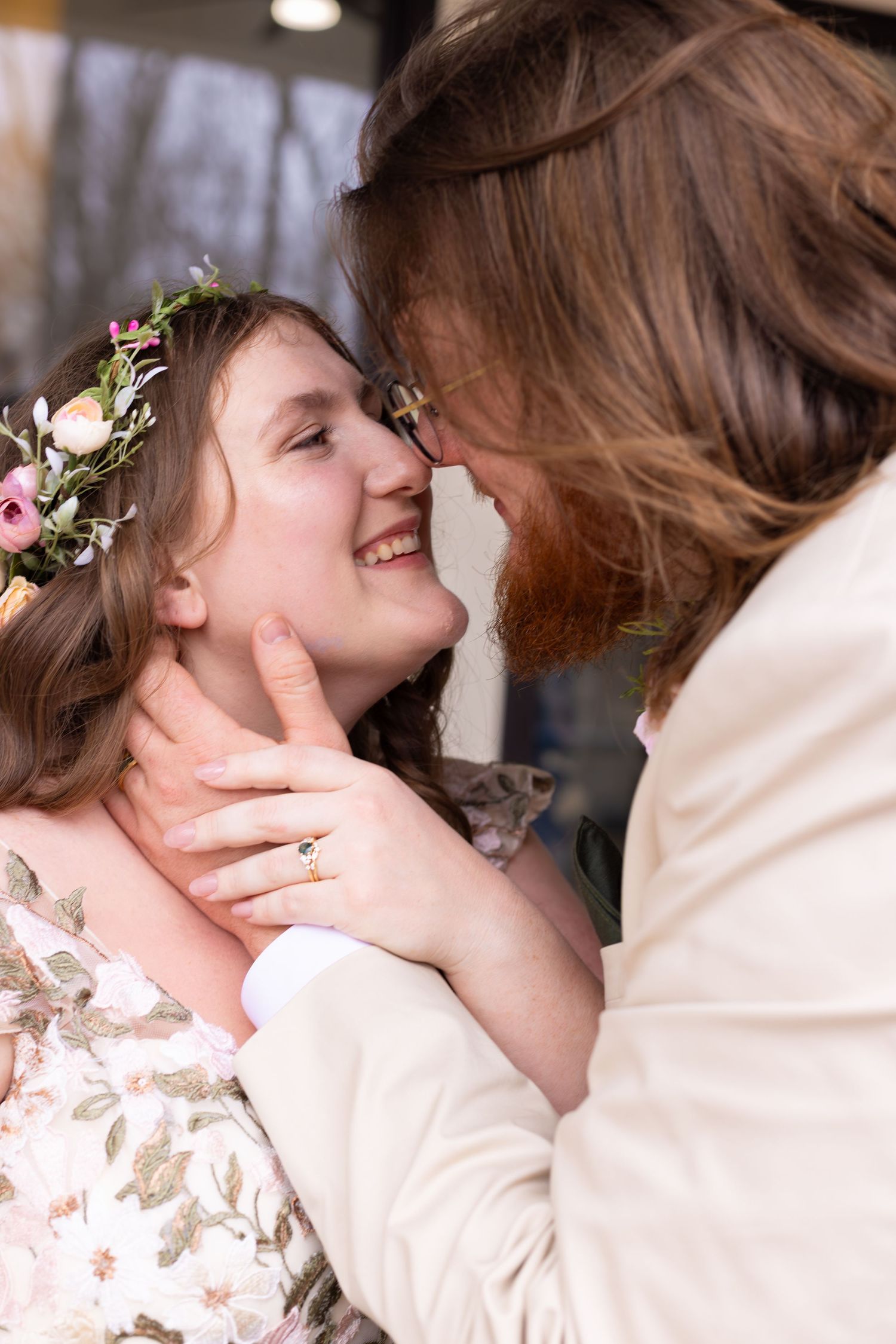 Two brides share a tender forehead touch, one wearing a floral crown and floral dress, the other in a cream suit.