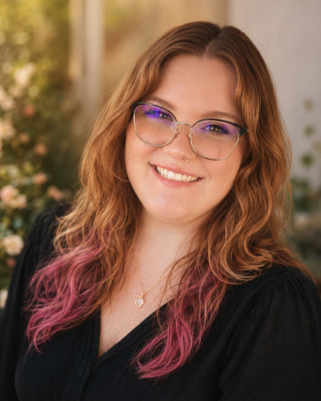Smiling young woman with red hair and pink highlights wearing glasses and a black top outdoors.