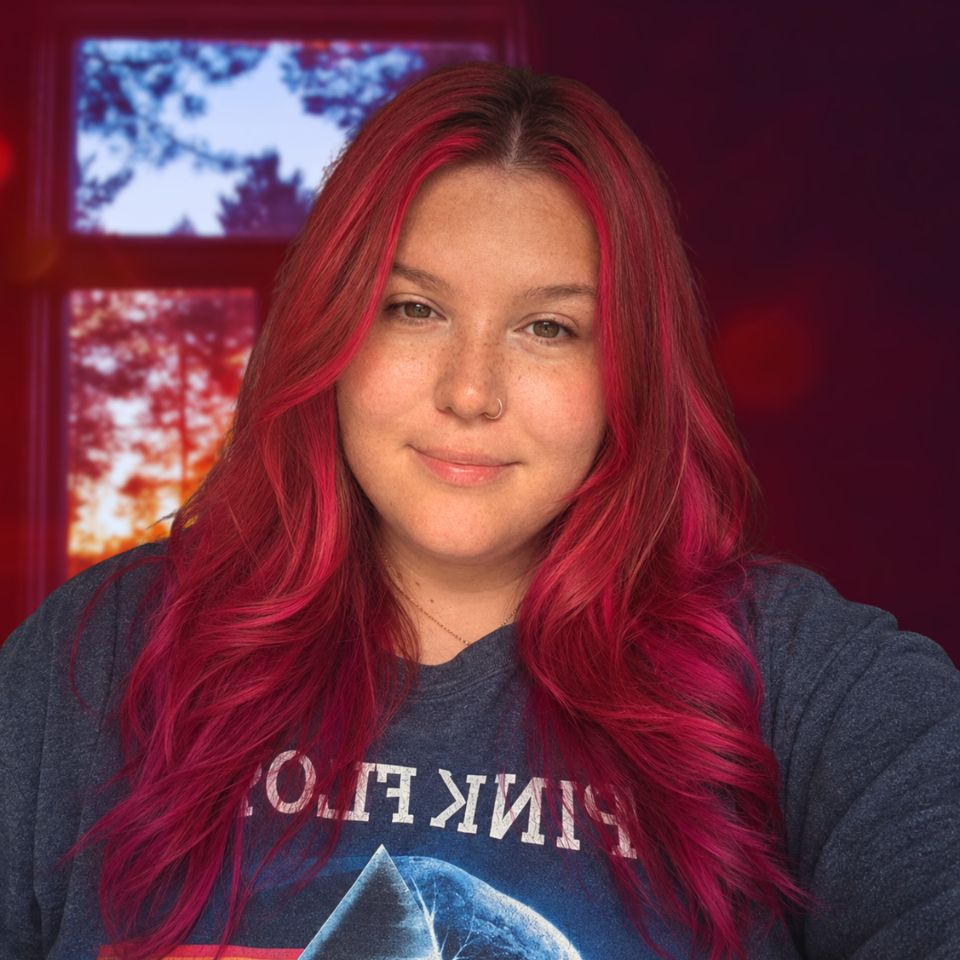 Young woman with vibrant red and pink wavy hair wearing a Pink Floyd t-shirt, posing indoors with warm red lighting.