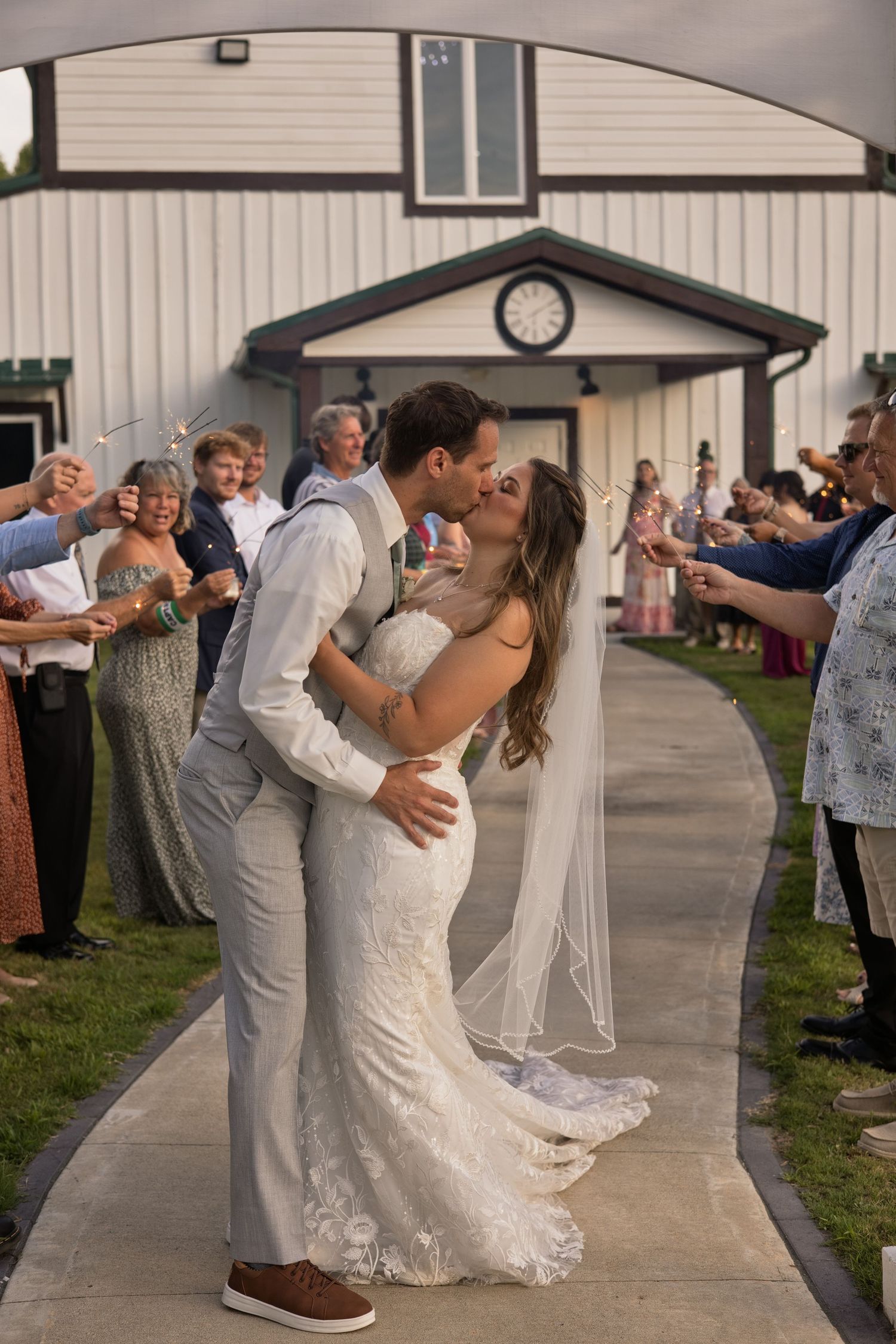 Newlywed couple sharing a kiss during sparkler send-off outside a white barn venue with guests cheering.