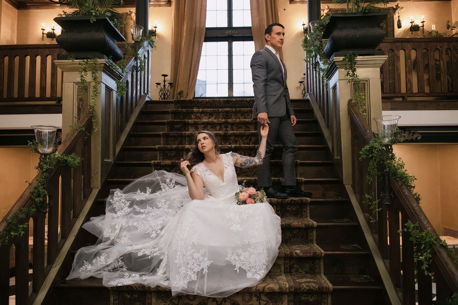 Bride in flowing white gown sits on ornate staircase while groom stands behind her, holding hands in elegant venue.