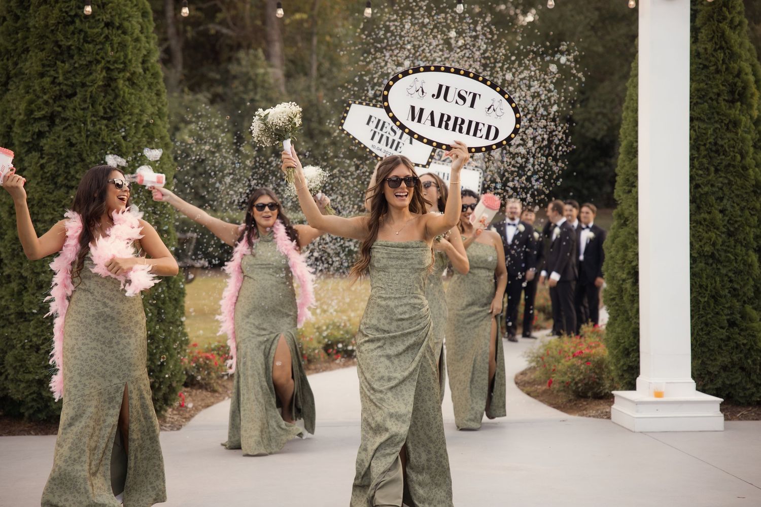 Bridesmaids in green gowns celebrate with confetti under a Just Married sign at outdoor wedding.