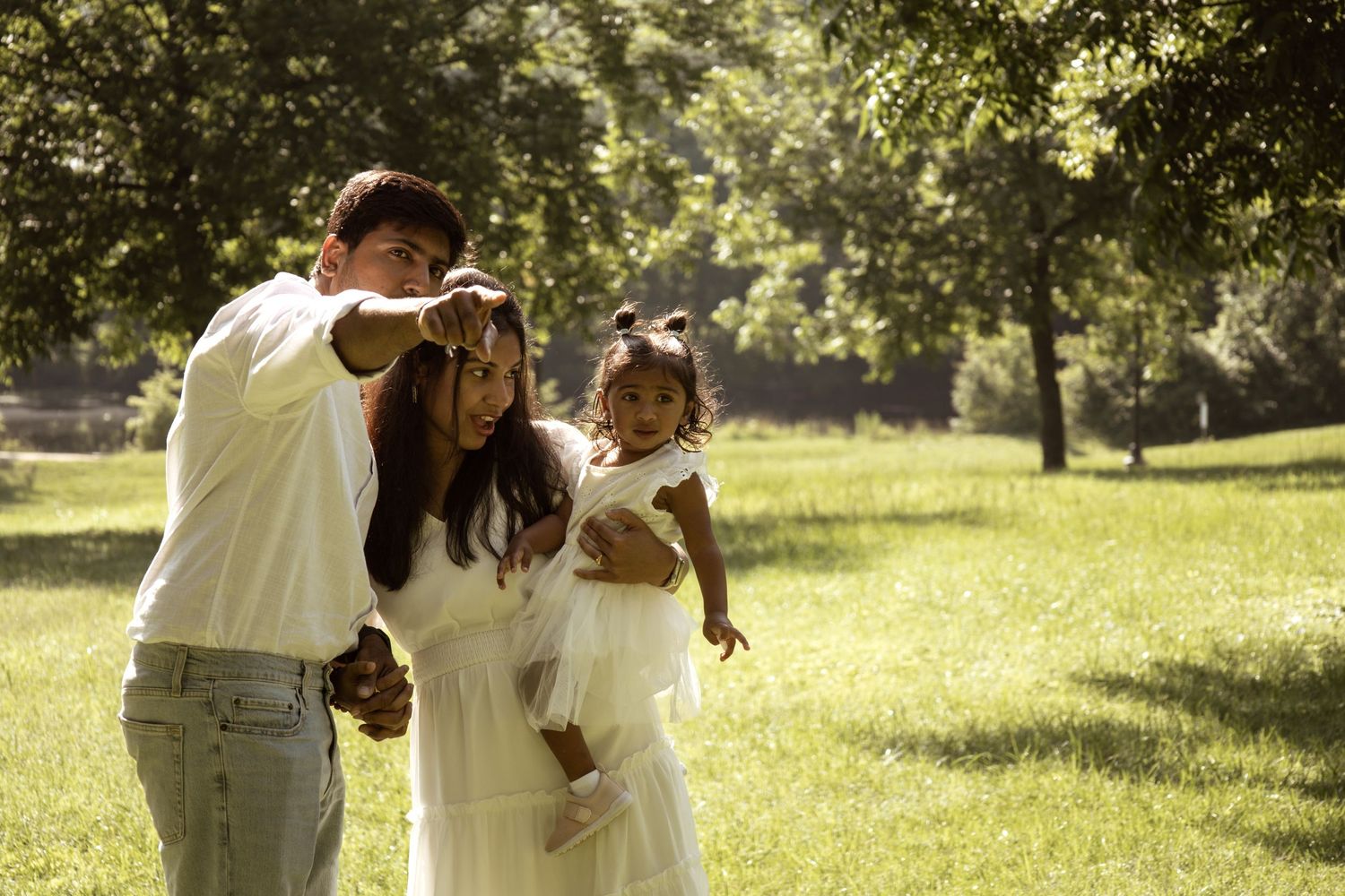 Pregnant woman in white dress with partner and toddler daughter enjoying a sunny day in a lush green park.