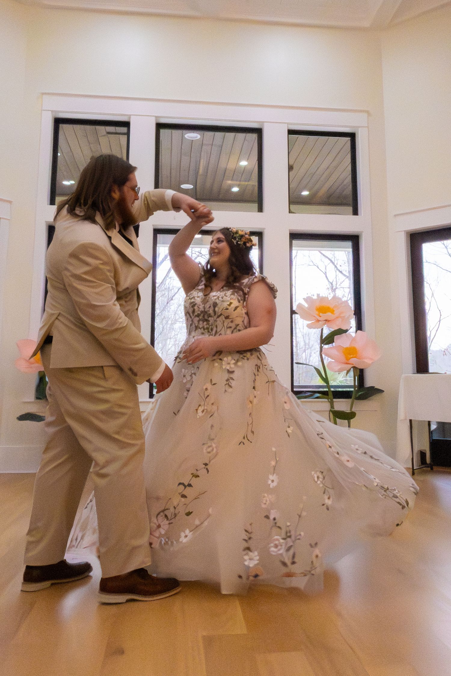 Bride in floral gown twirls with groom in tan suit during first dance at elegant indoor wedding venue.