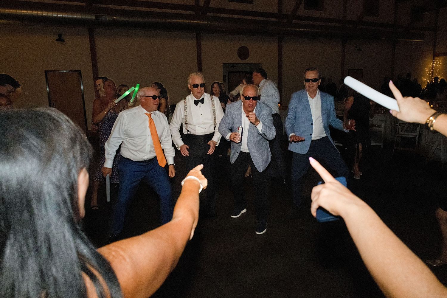 Group of men dancing at a wedding reception party, wearing sunglasses and formal attire, holding glow sticks.