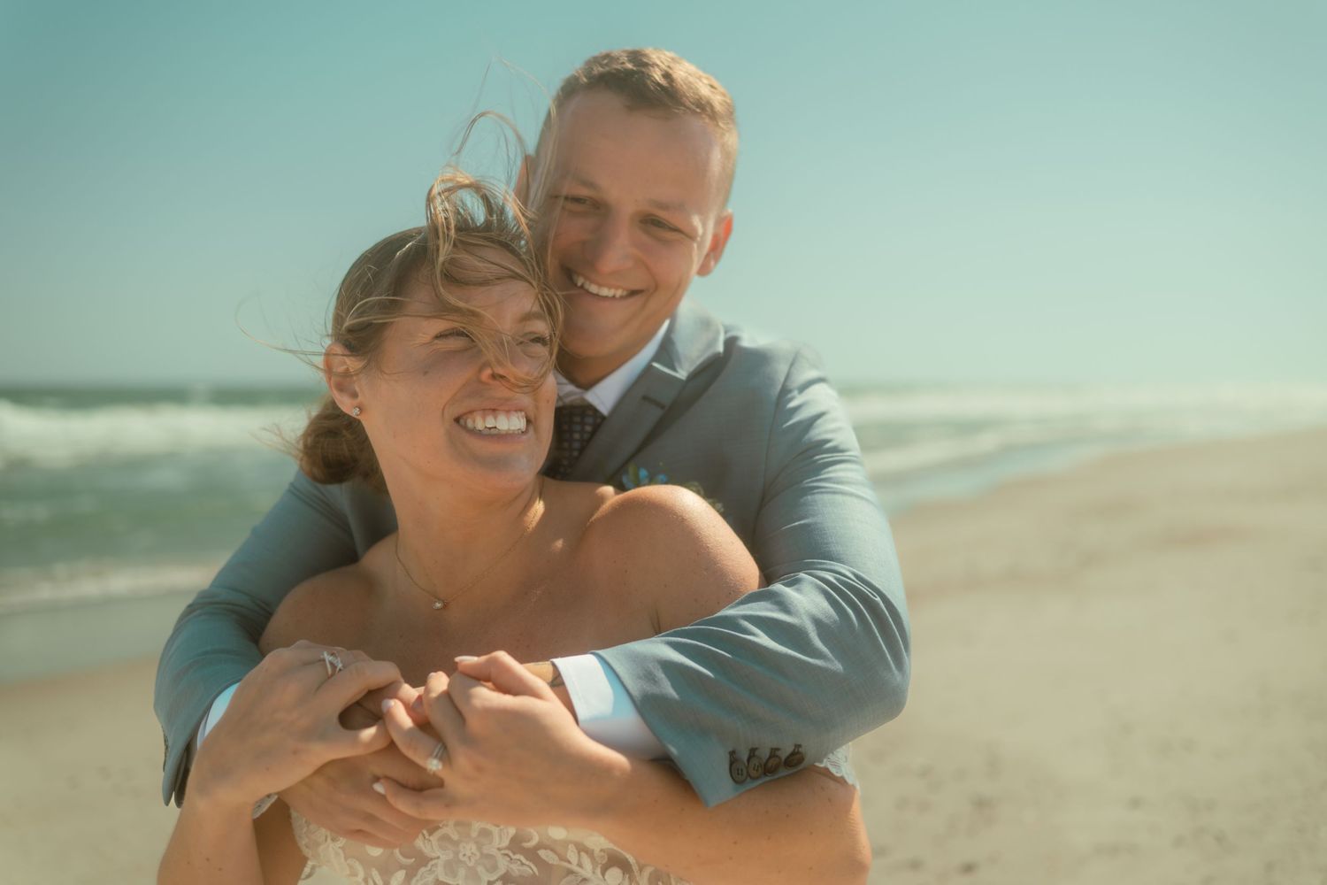 Happy couple embracing on a sunny beach in wedding attire, smiling joyfully.