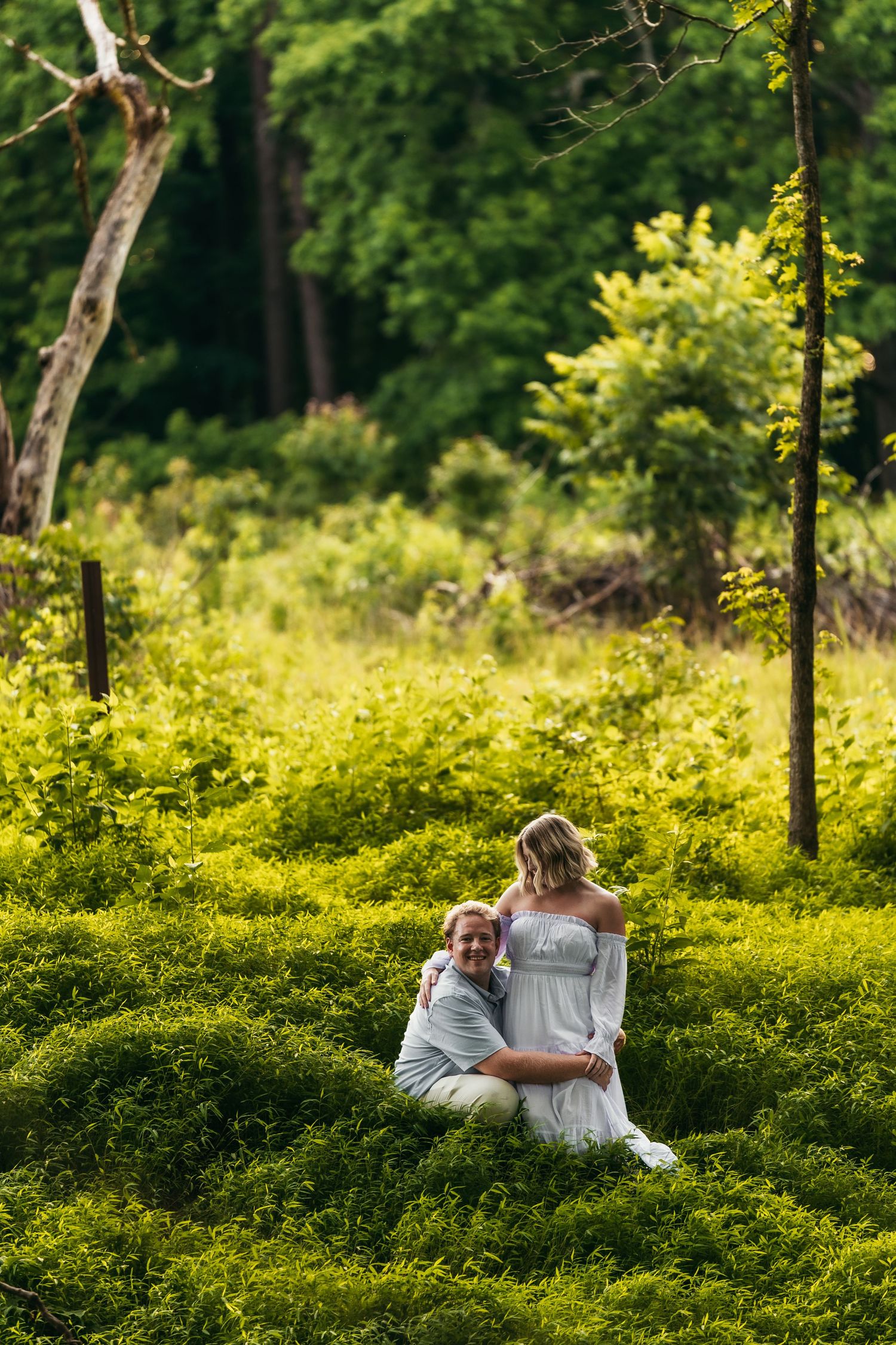 Couple embracing in lush green meadow surrounded by trees, woman in white off-shoulder dress, outdoor portrait photography.