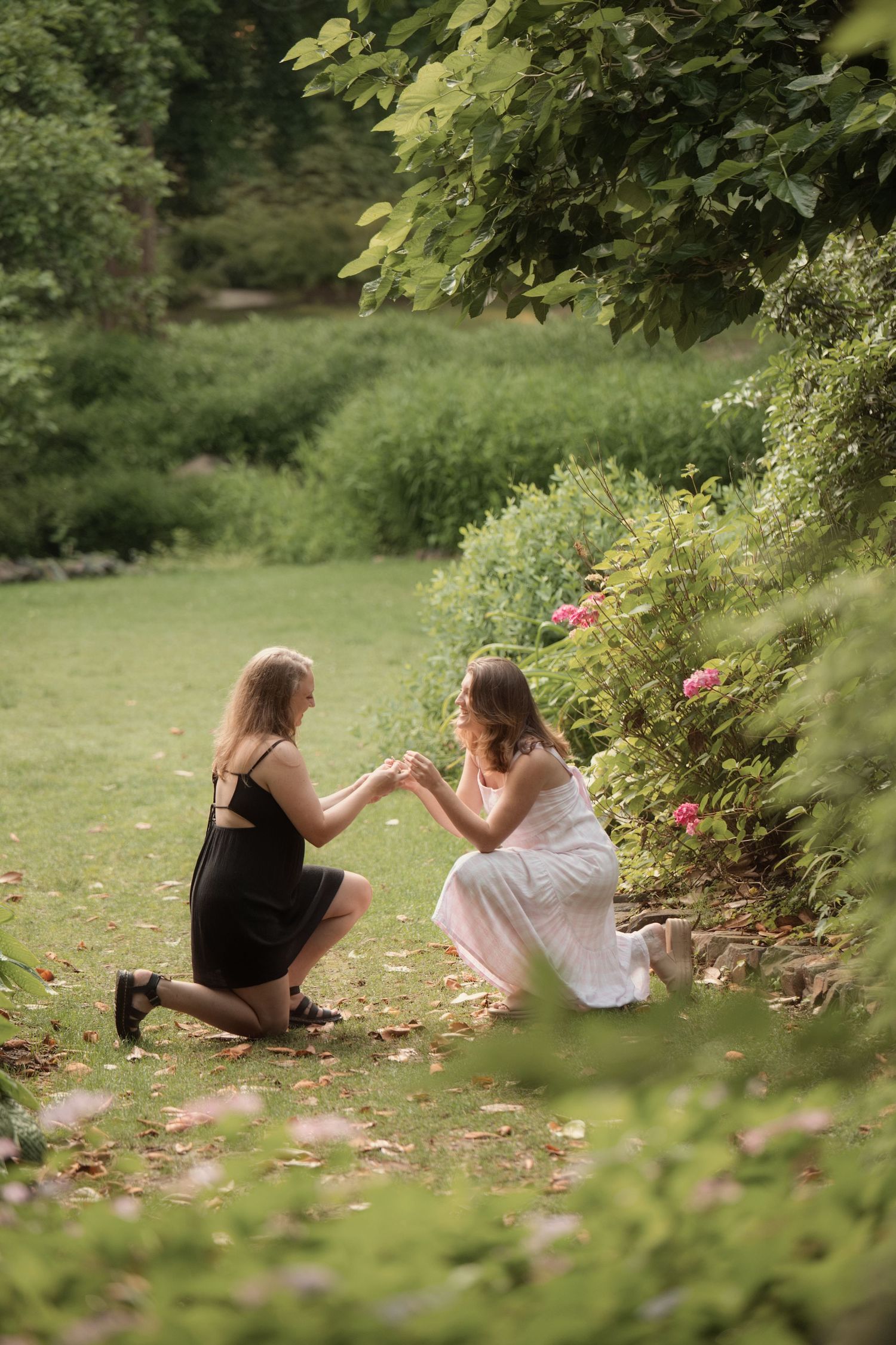 Two women share a romantic marriage proposal in a lush green garden, one kneeling in a black dress offering a ring.