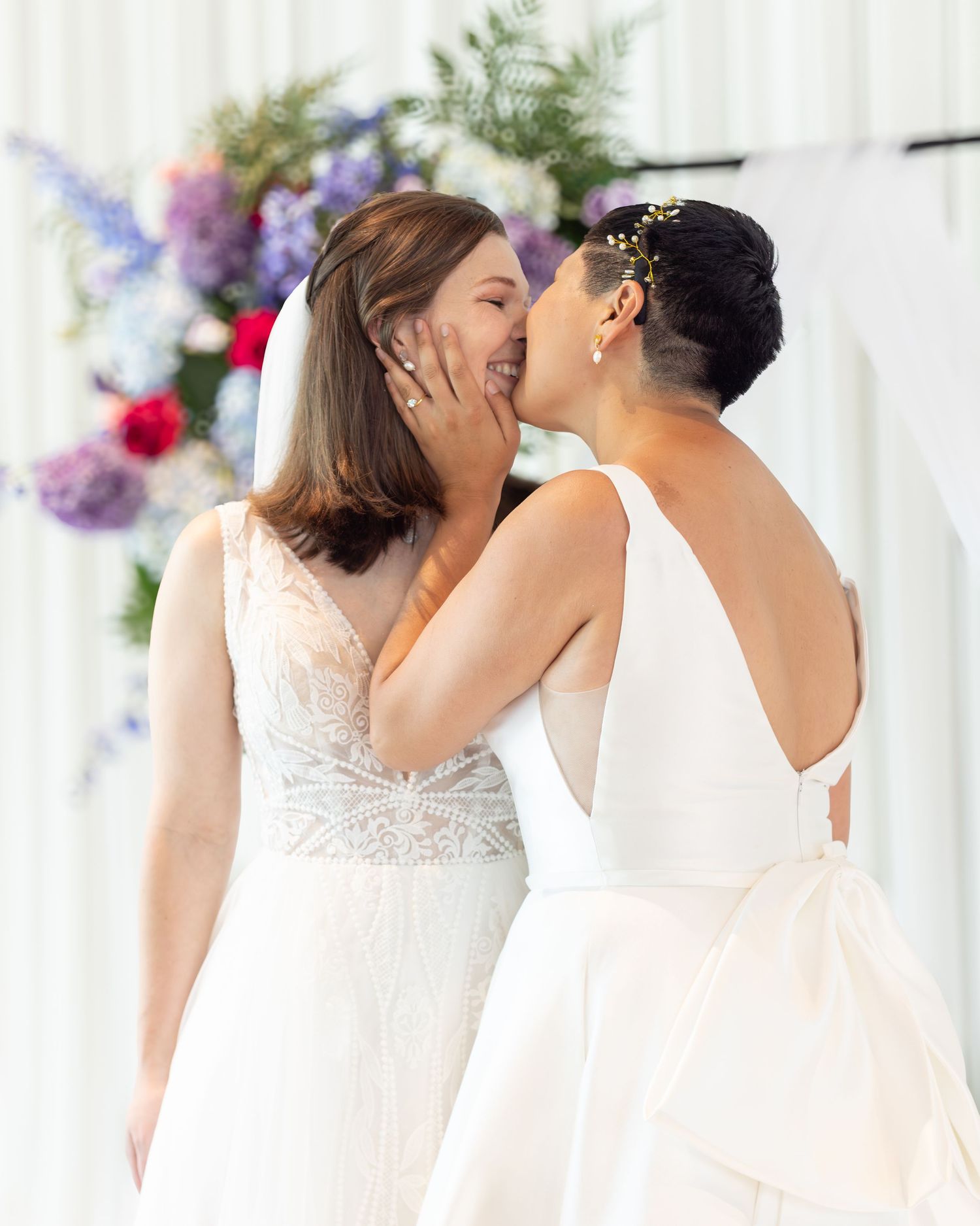 Two brides in white wedding gowns share a kiss at their outdoor ceremony with colorful floral backdrop.