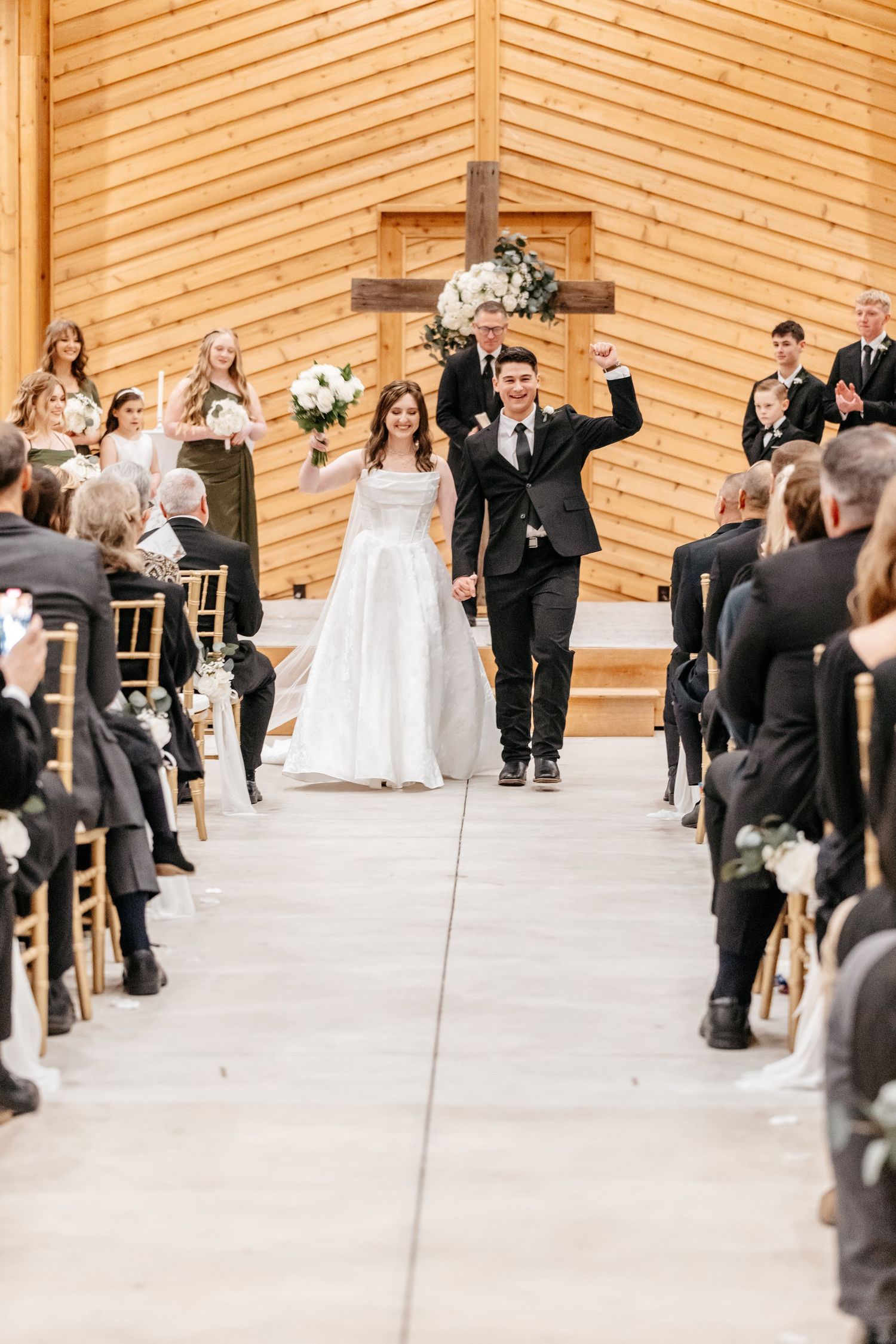 Joyful newlyweds walk down the aisle in a rustic wooden chapel, groom raising fist in celebration.