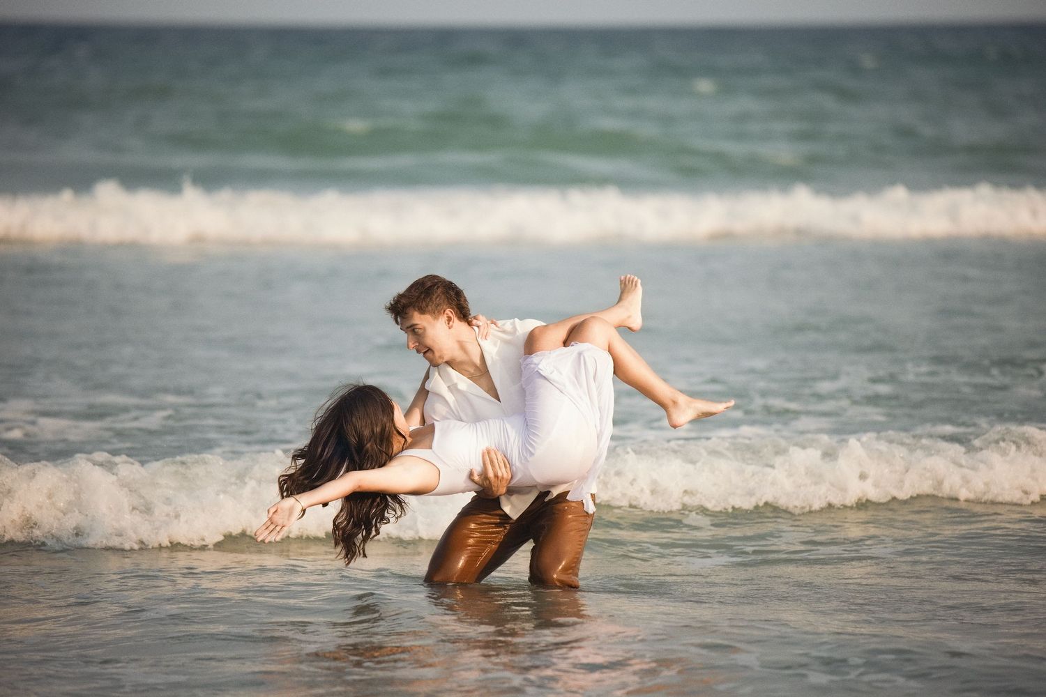 Couple playing in ocean waves on a sandy beach, man lifting woman in white dress while waves crash around them.