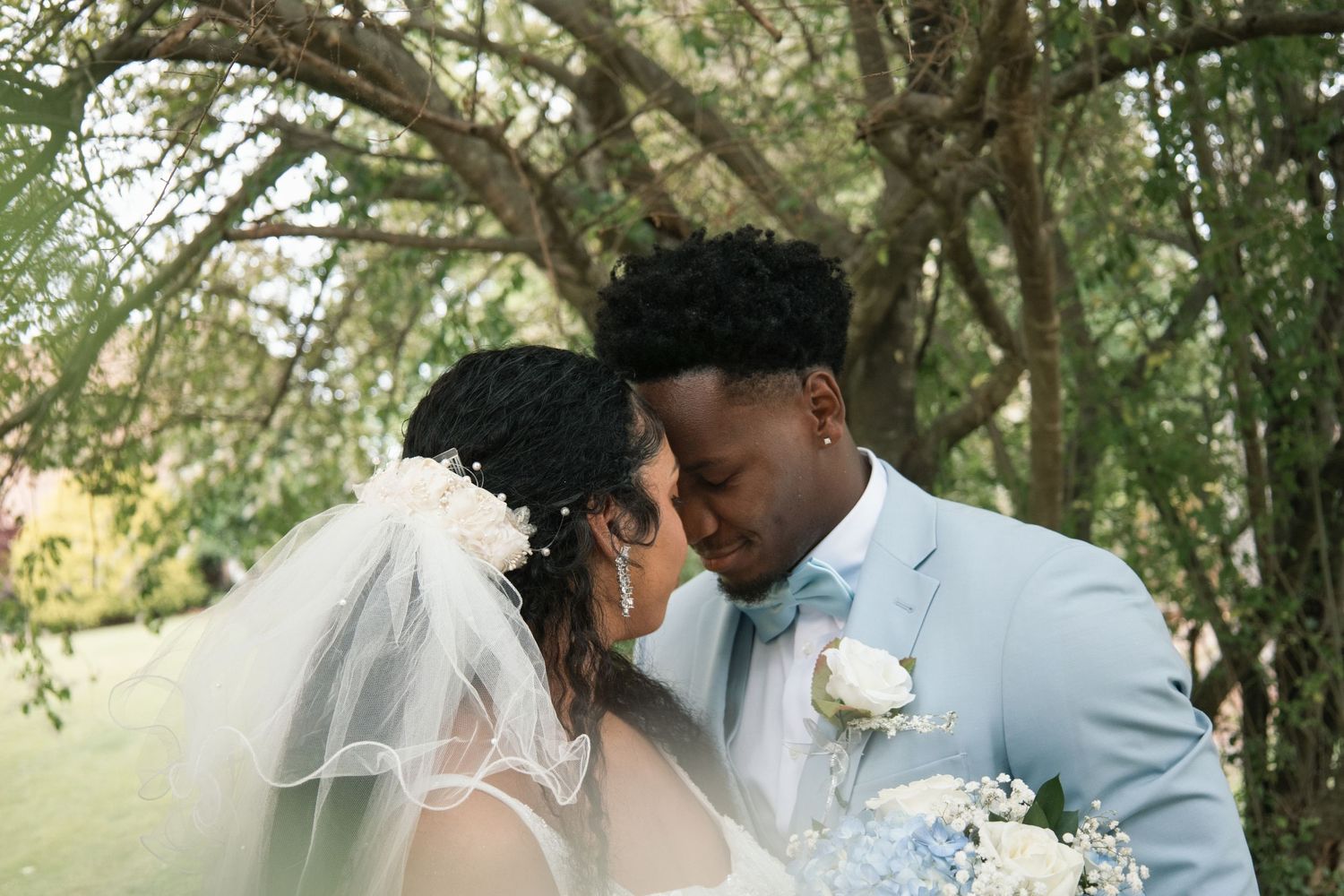 A newlywed couple shares a romantic forehead touch outdoors, bride in white veil and groom in light blue suit.