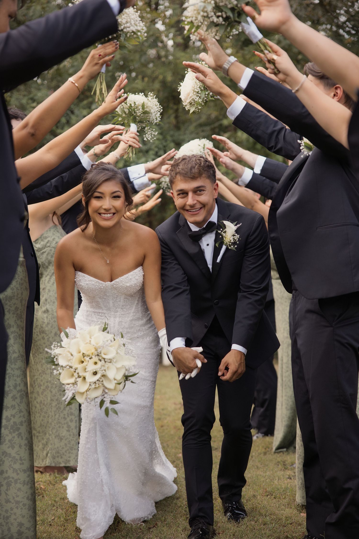 Bride and groom walk through an arch of raised hands and flowers formed by wedding guests outdoors.