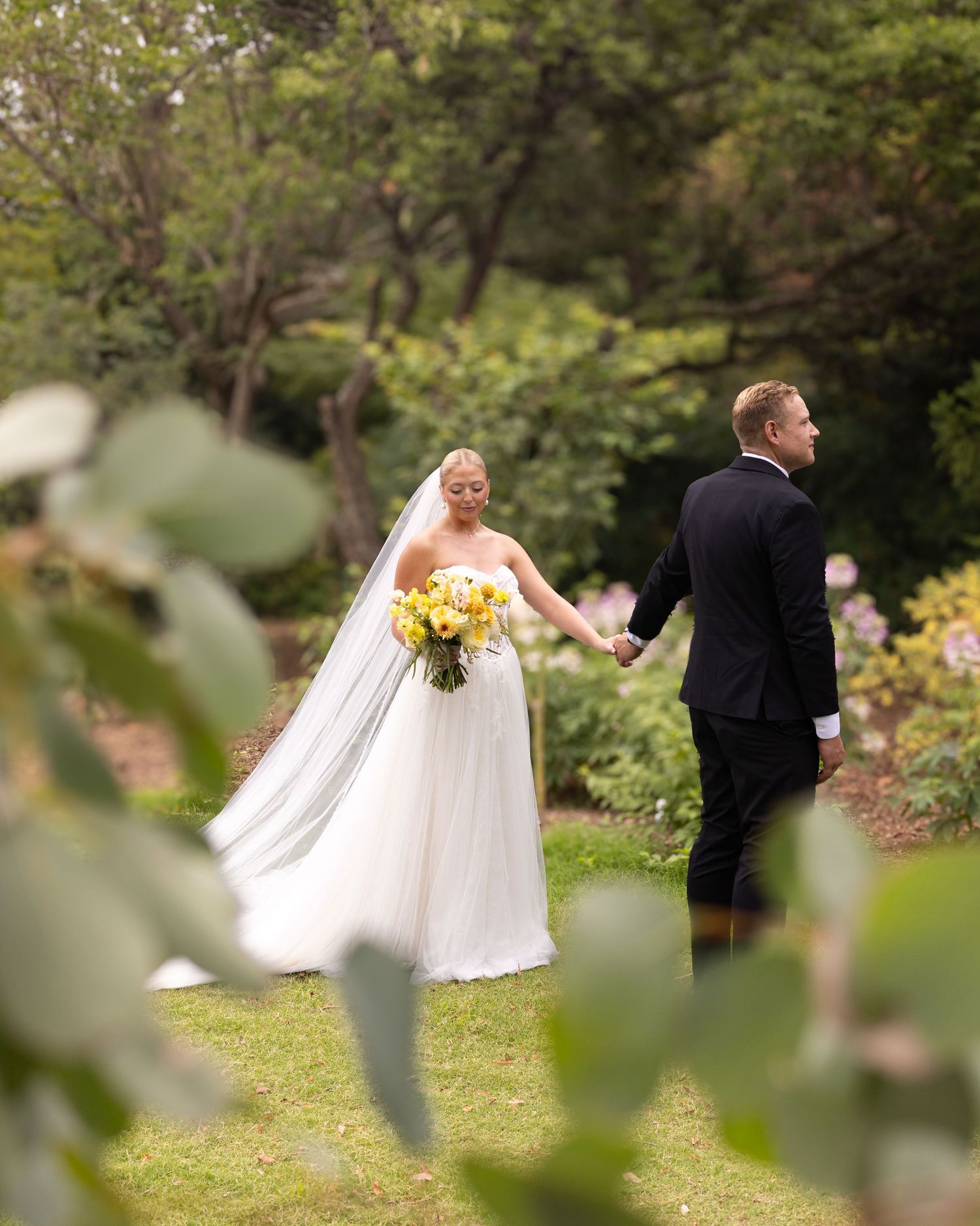 Bride in white gown holding yellow bouquet walks with groom in black suit through lush garden on wedding day.