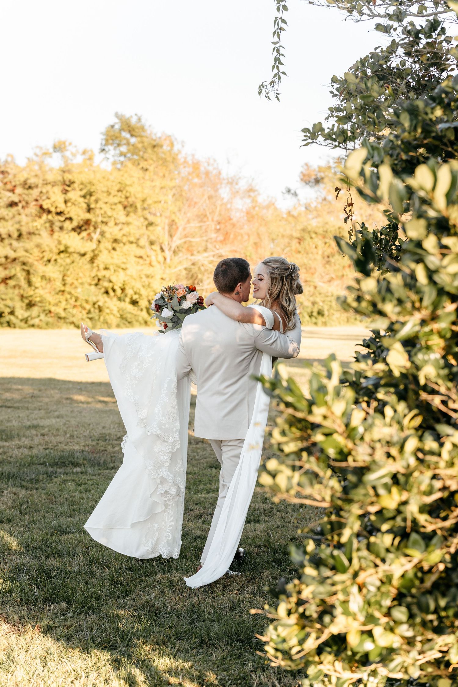 Groom lifting bride in white lace dress outdoors, sharing a romantic kiss surrounded by lush greenery on their wedding day.