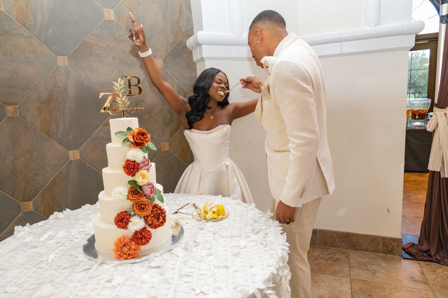 A bride and groom celebrate joyfully while cutting their elegant white wedding cake adorned with colorful fall flowers.
