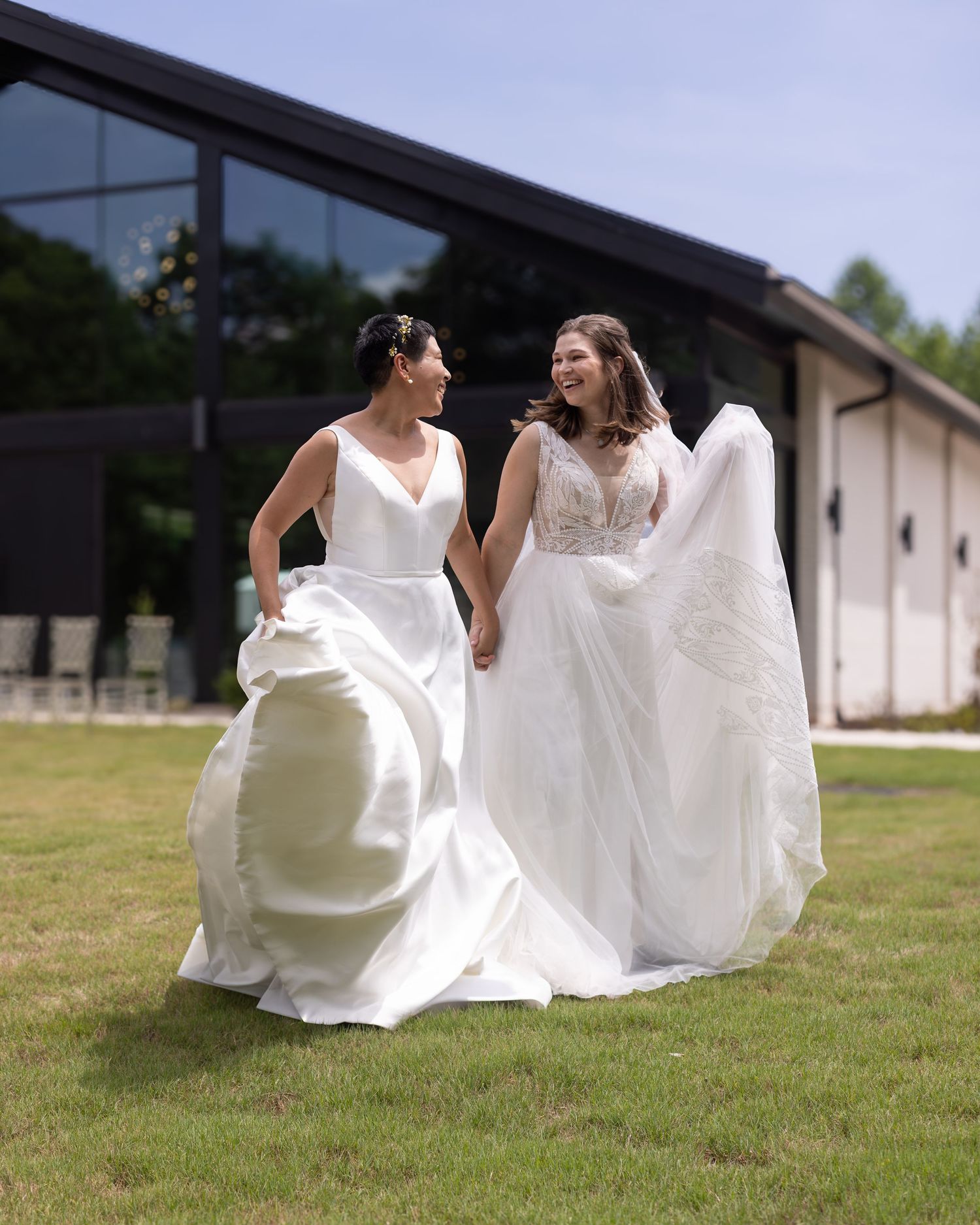 Two brides in white wedding gowns hold hands and walk joyfully across green lawn outside modern venue.