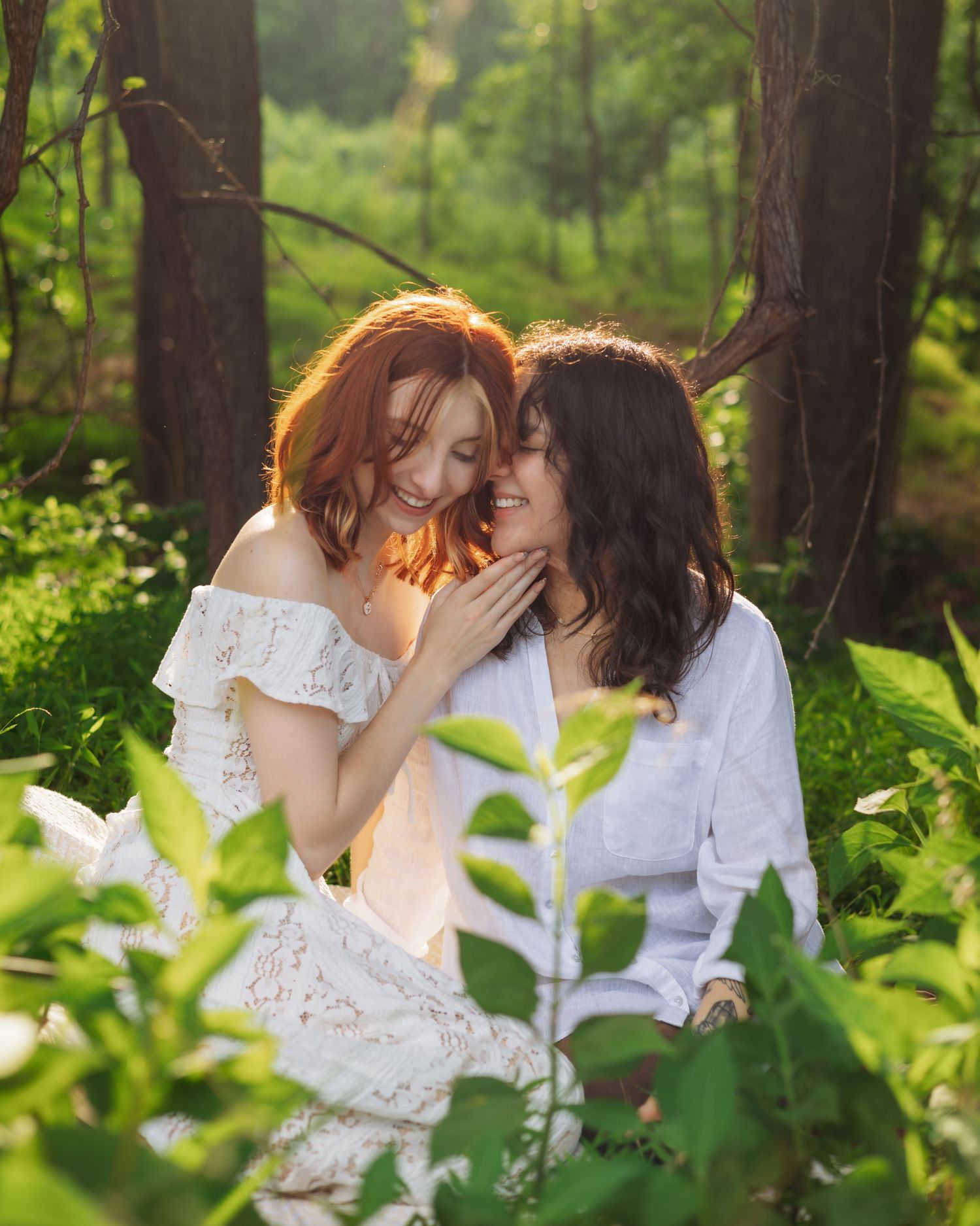 Two young women in white outfits share a tender moment sitting together in a sunlit forest surrounded by lush green foliage.