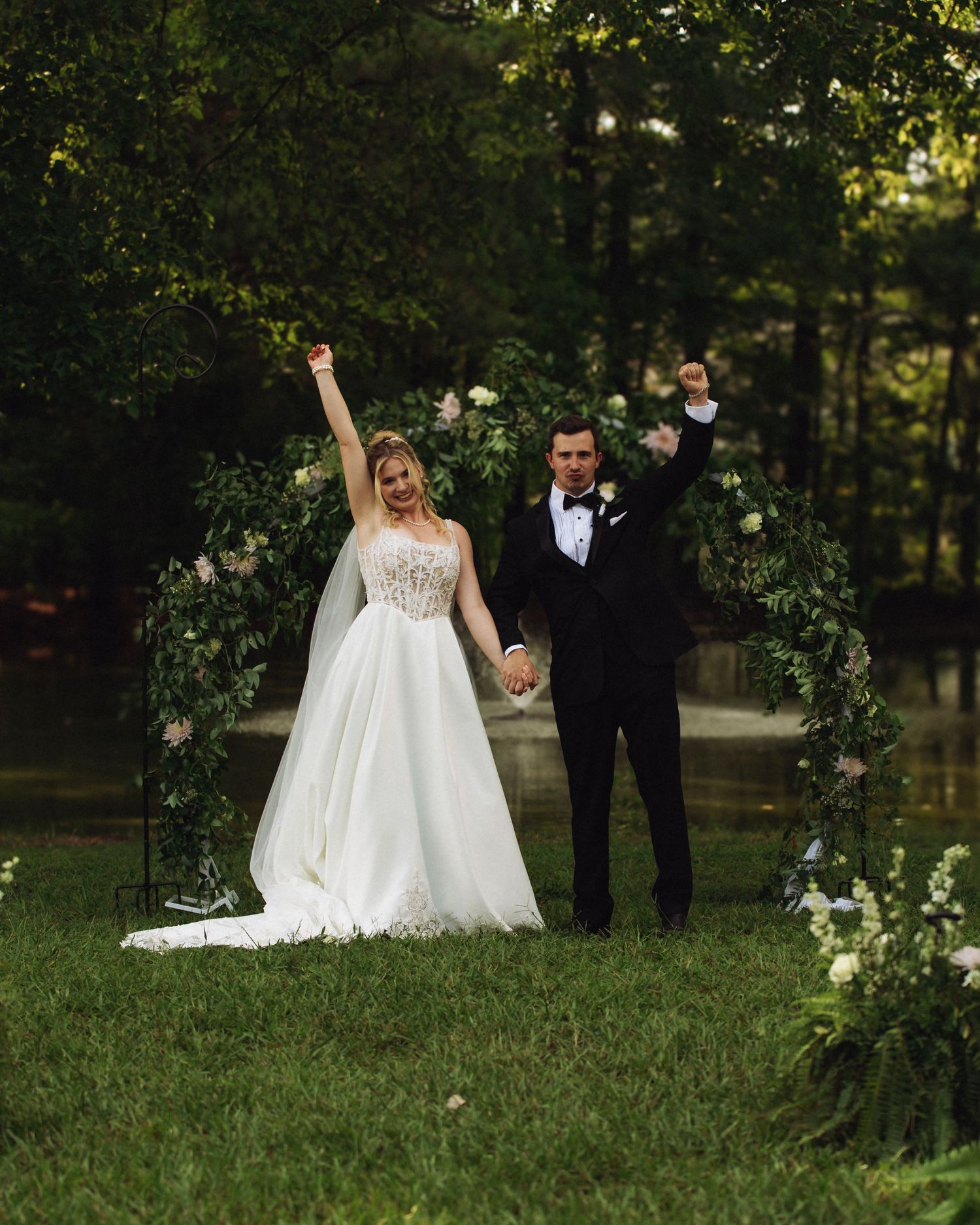 Joyful bride and groom celebrate under floral arch outdoors, raising arms in triumph on lush green lawn.