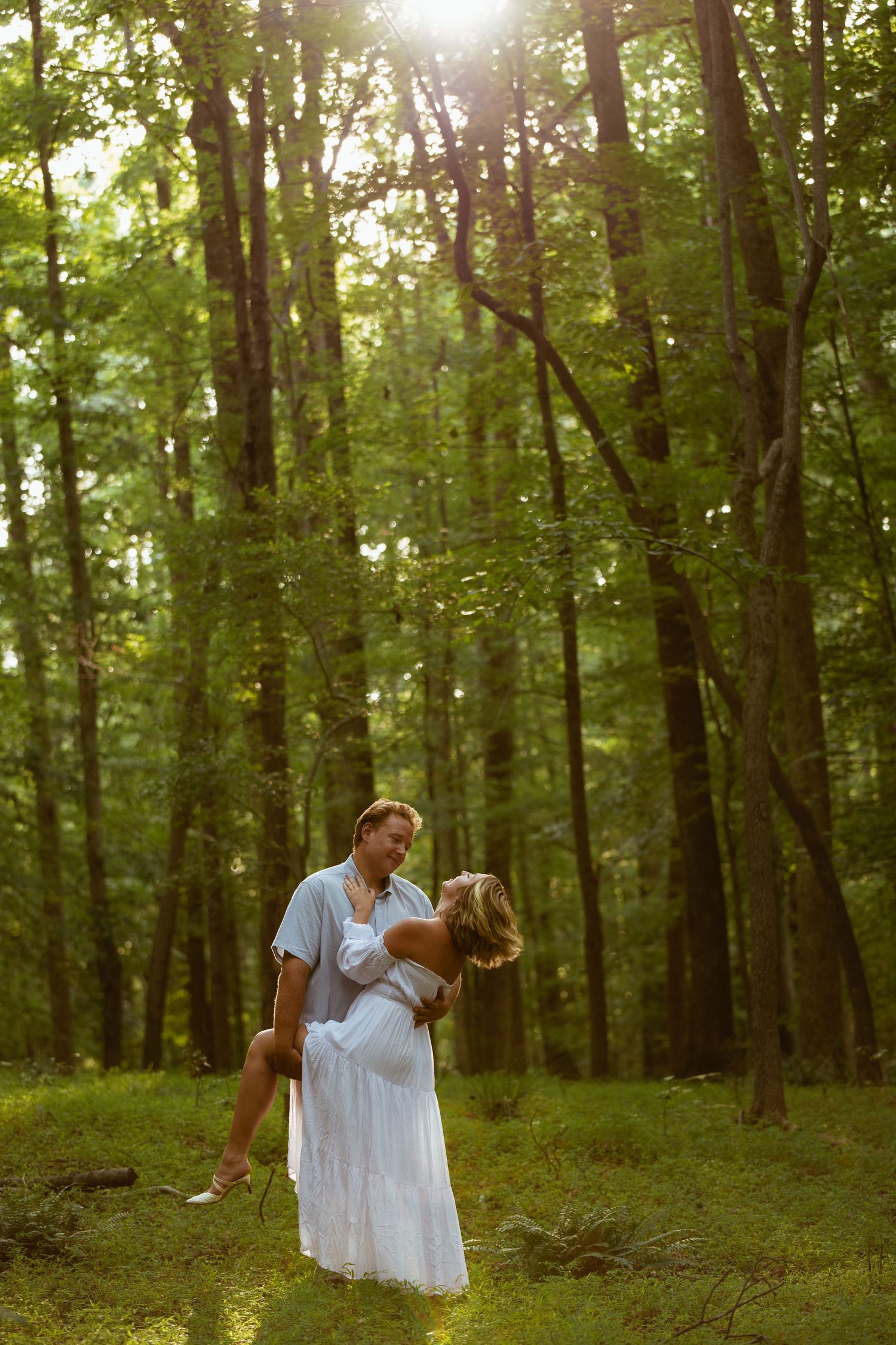 A couple embraces in a sunlit forest, man lifting pregnant woman in white dress among tall green trees.