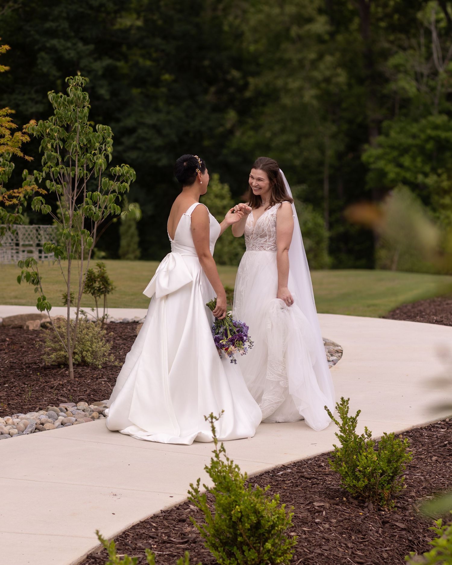 Two brides in white wedding gowns holding bouquets share a joyful moment on a garden pathway surrounded by lush greenery.