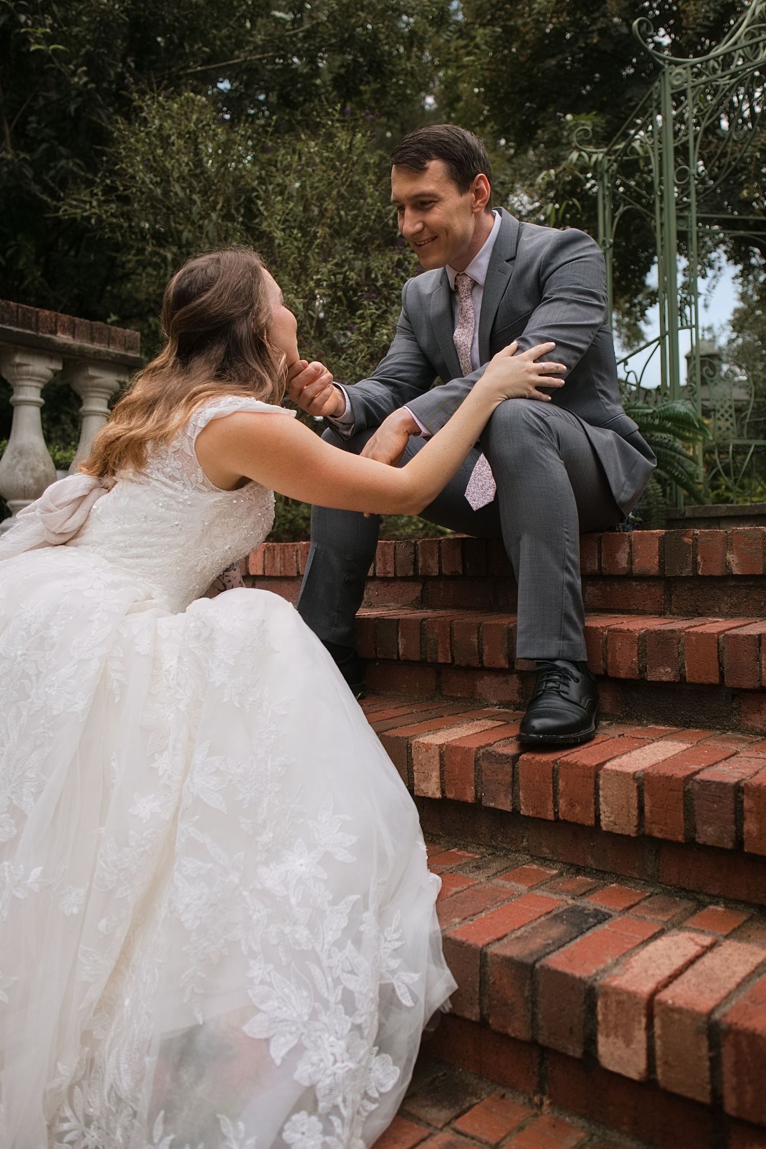 Bride in lace gown and groom in gray suit share a tender moment on brick garden steps.