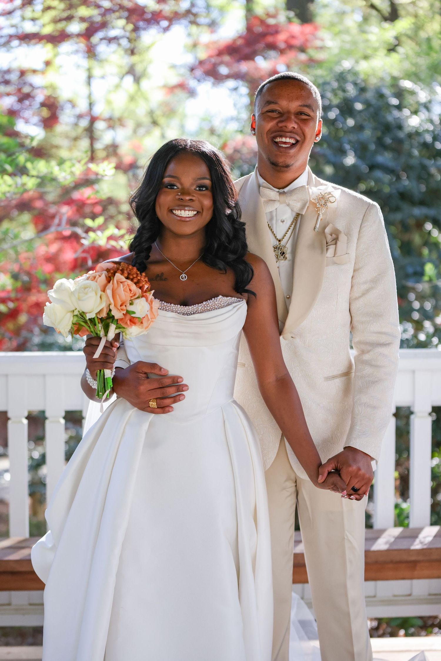Happy newlyweds pose outdoors, bride in white gown holding peach bouquet, groom in cream suit, surrounded by autumn foliage.