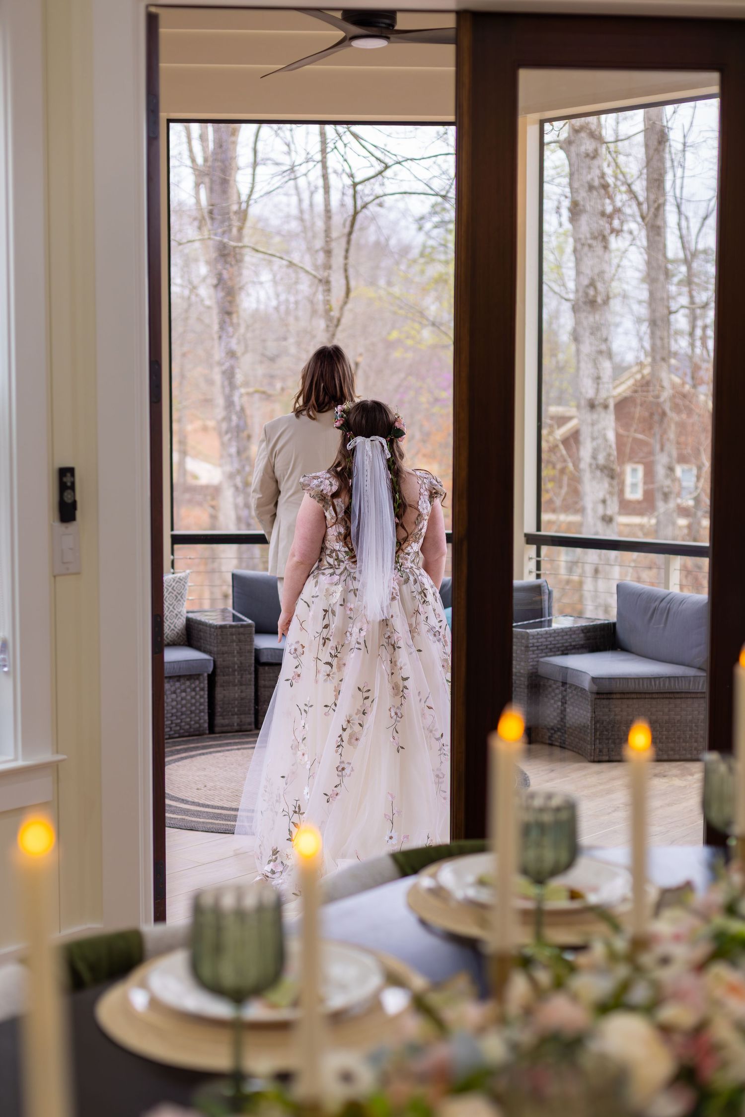 Bride in floral gown with veil stands in doorway with groom, overlooking wooded deck, candles and table setting in foreground.