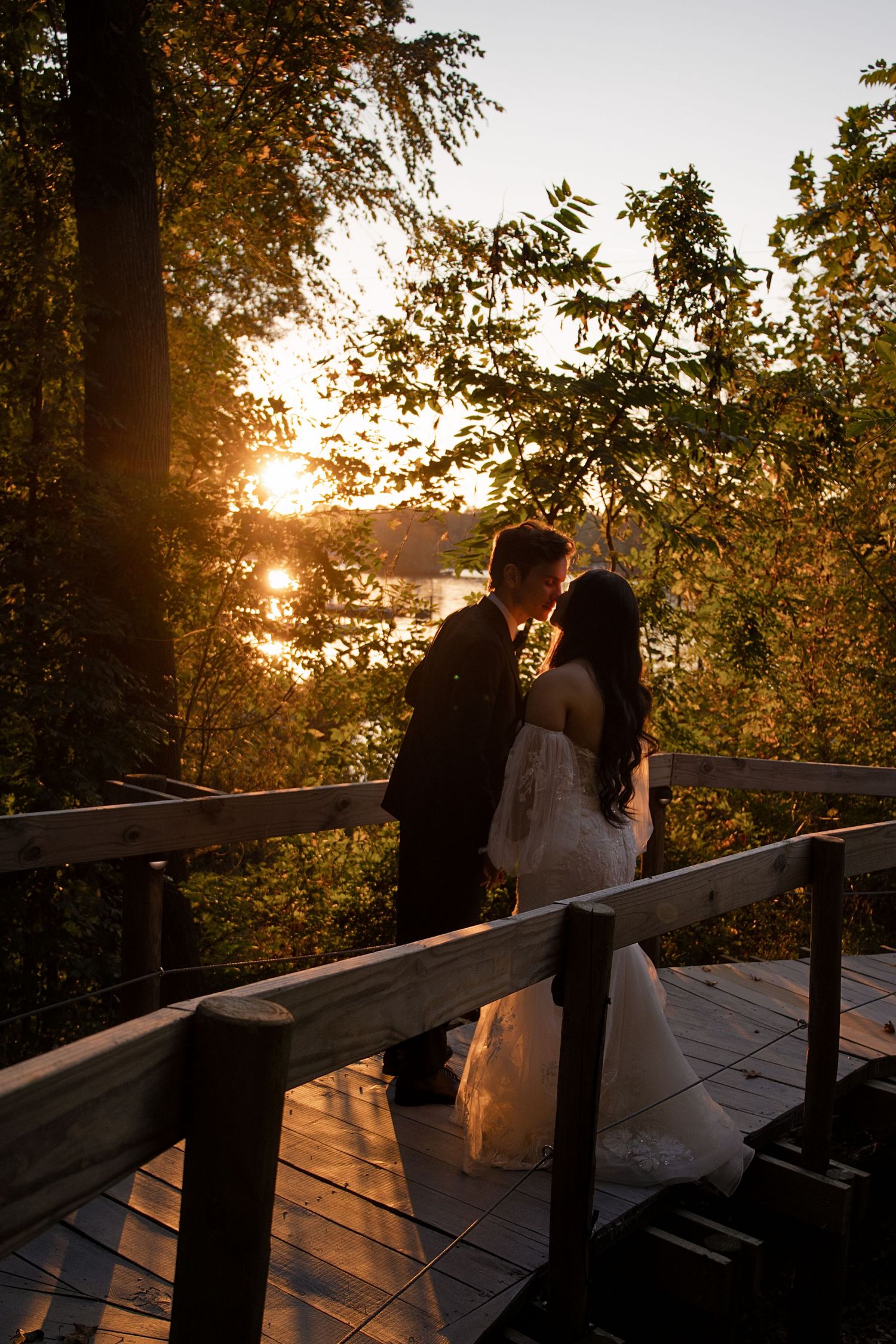 Bride and groom kissing on a wooden deck at sunset, surrounded by autumn trees and a shimmering lake backdrop.