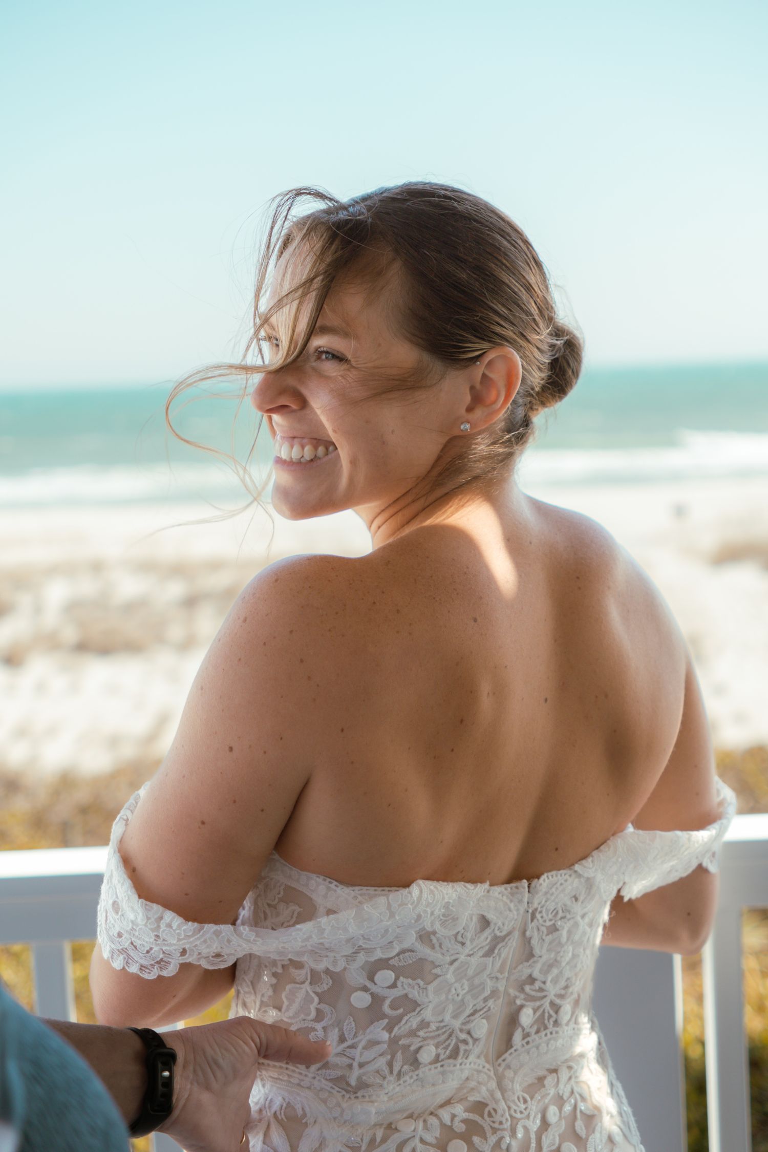 Smiling bride in off-shoulder lace wedding dress on beach deck with ocean view on sunny day.