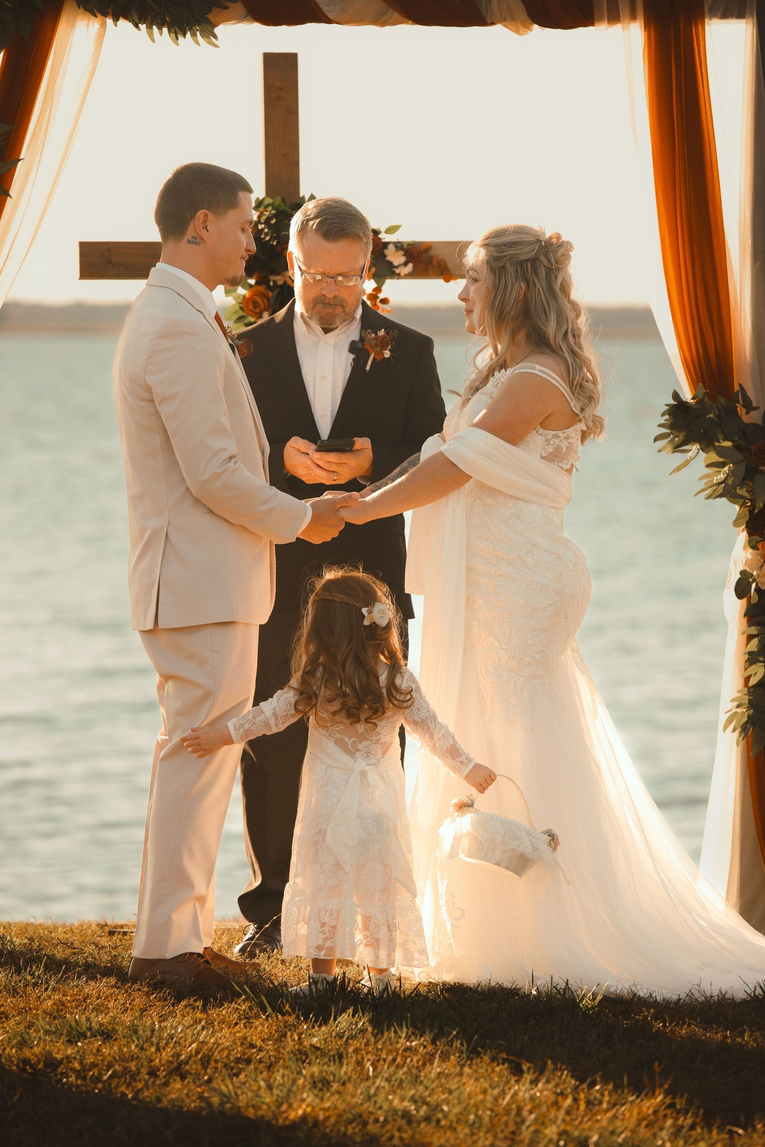 Bride and groom exchange vows at waterfront wedding ceremony with flower girl and officiant under wooden cross altar.