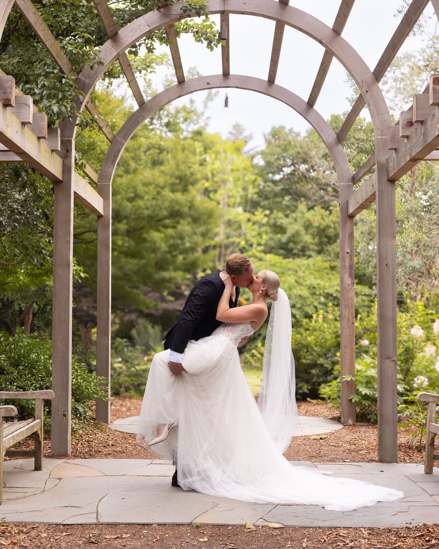 A bride and groom share a romantic dip kiss under a wooden garden arch surrounded by lush greenery.