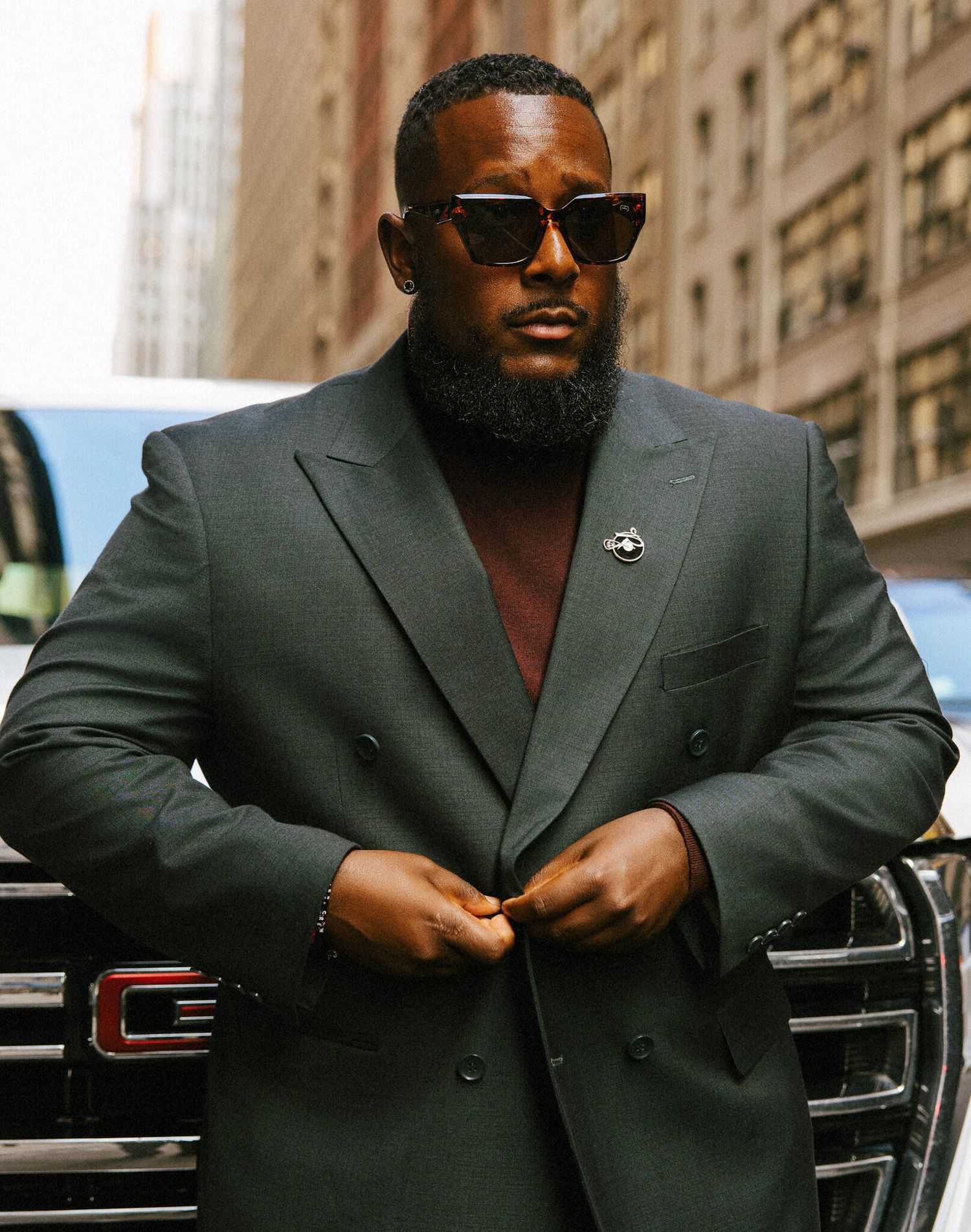 A well-dressed man in a dark suit and sunglasses poses confidently against an urban backdrop.