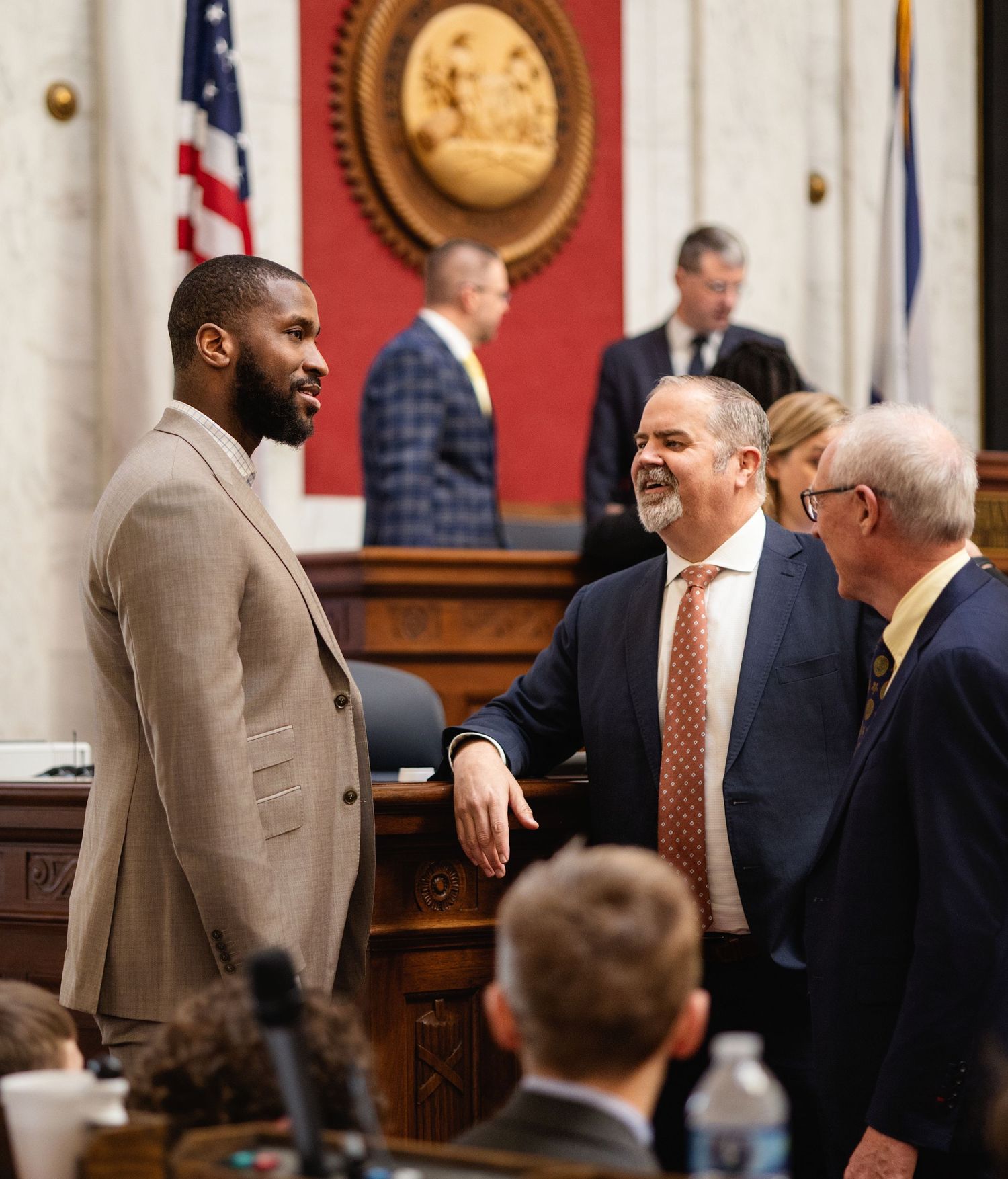 People in formal attire gather and converse during a legislative session in a government chamber.