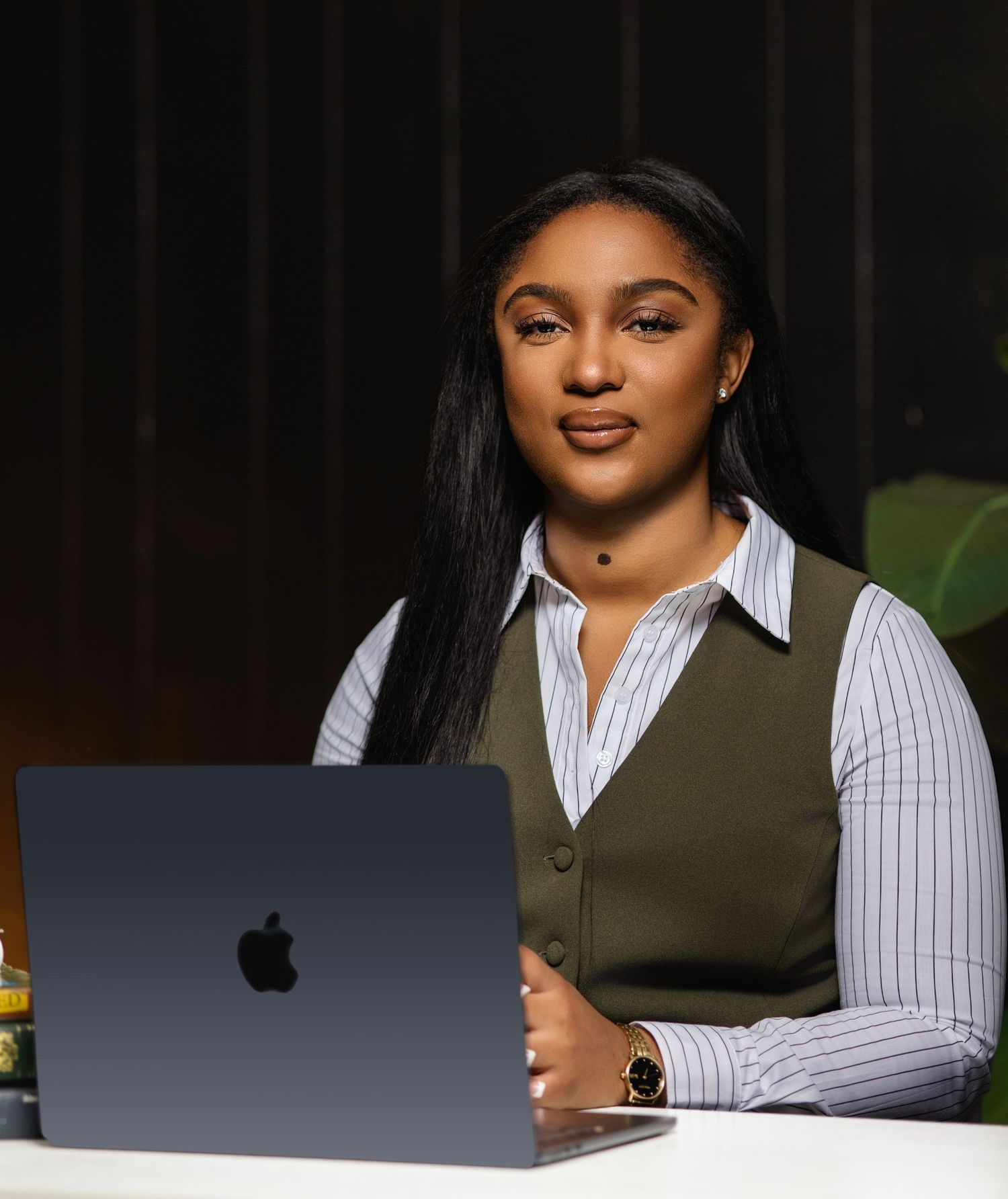 Professional in a striped shirt and olive vest sits at desk working on MacBook laptop in dark studio setting.