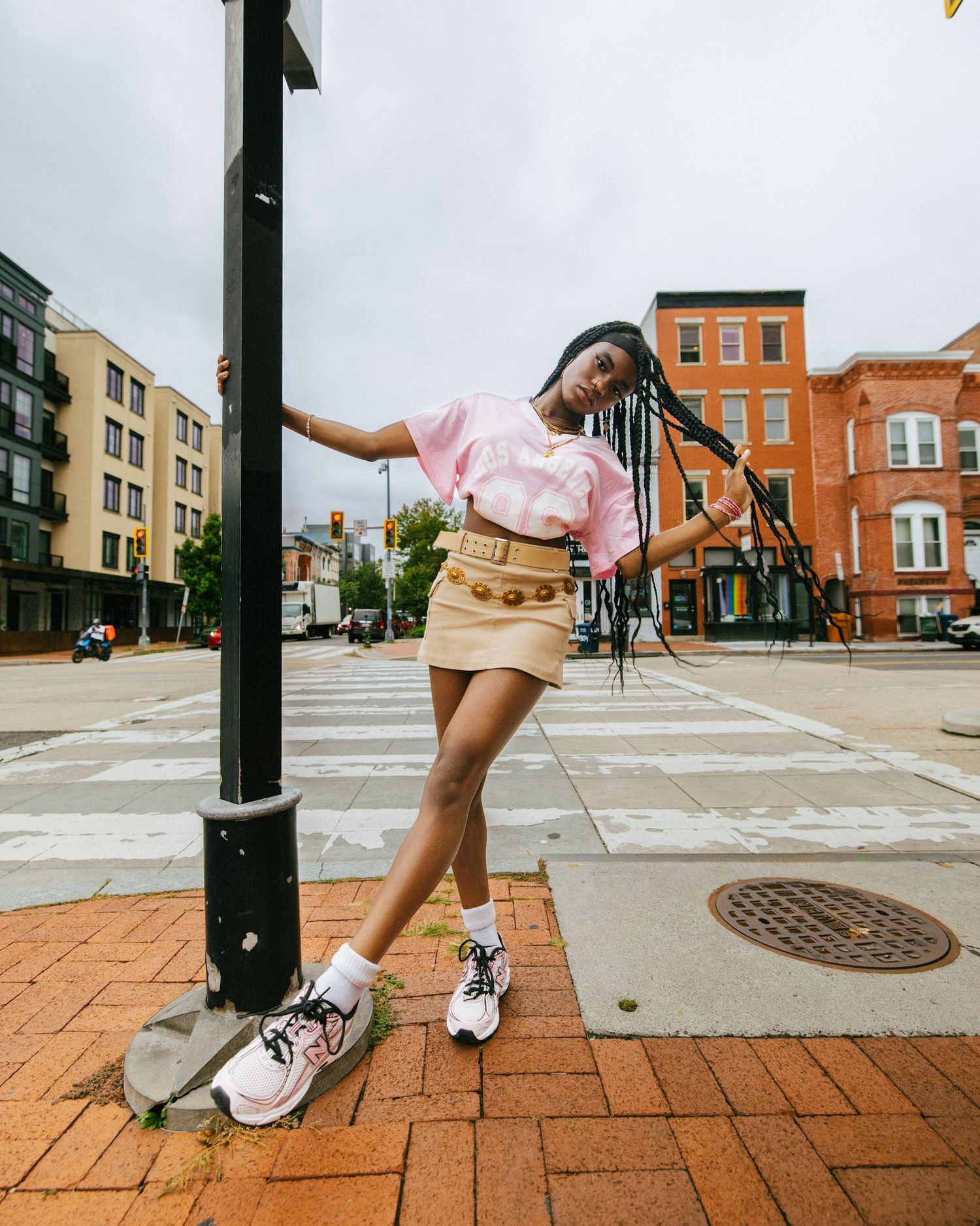 Model in pink top and khaki skirt poses playfully against lamppost on brick sidewalk in urban setting.