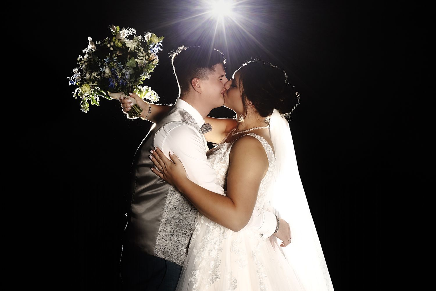 A romantic wedding couple shares a kiss at night with a bright flash illuminating their silhouette against a dark background.