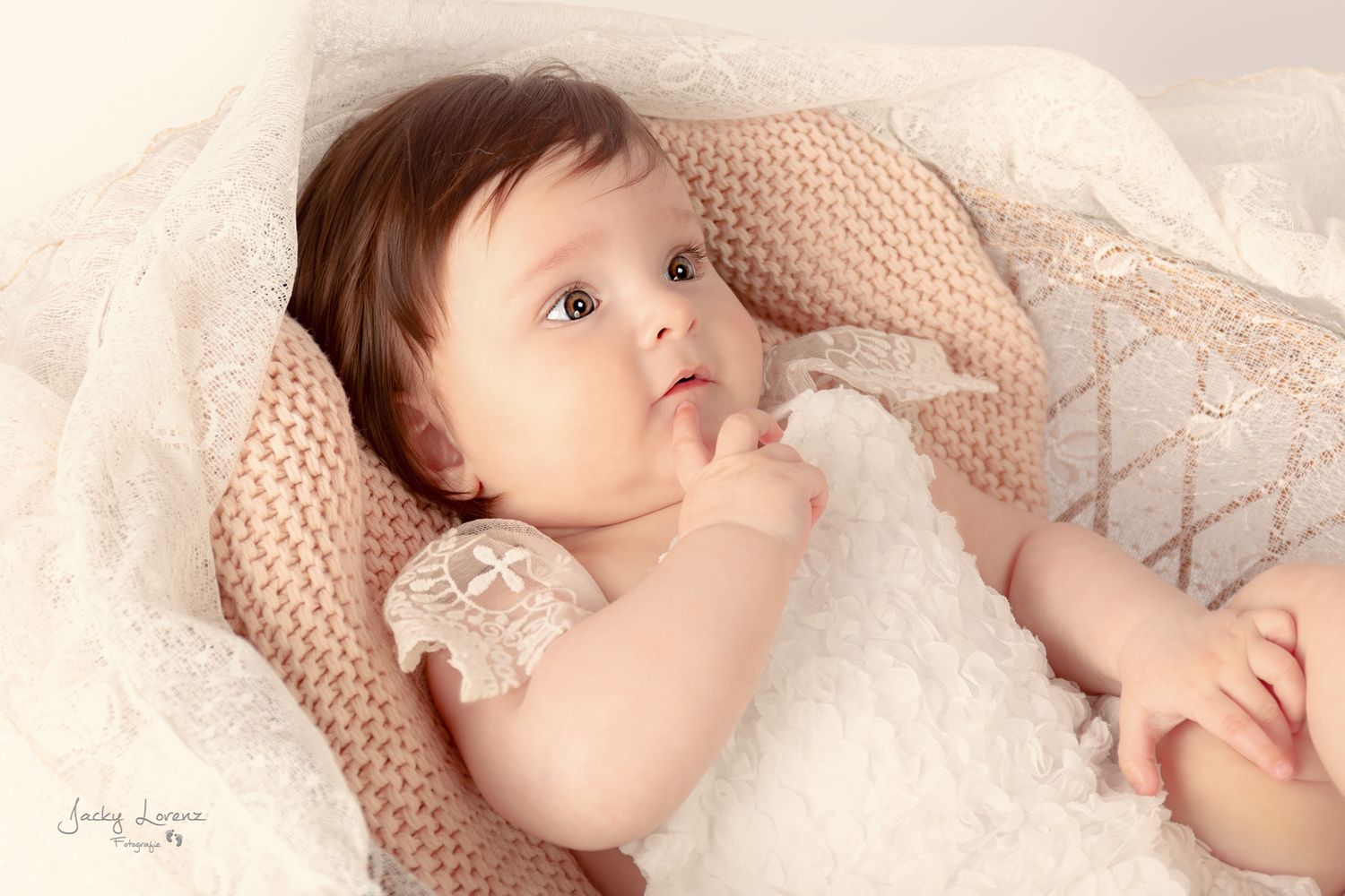 A sleeping baby in white lace clothing lies peacefully on soft pink bedding.