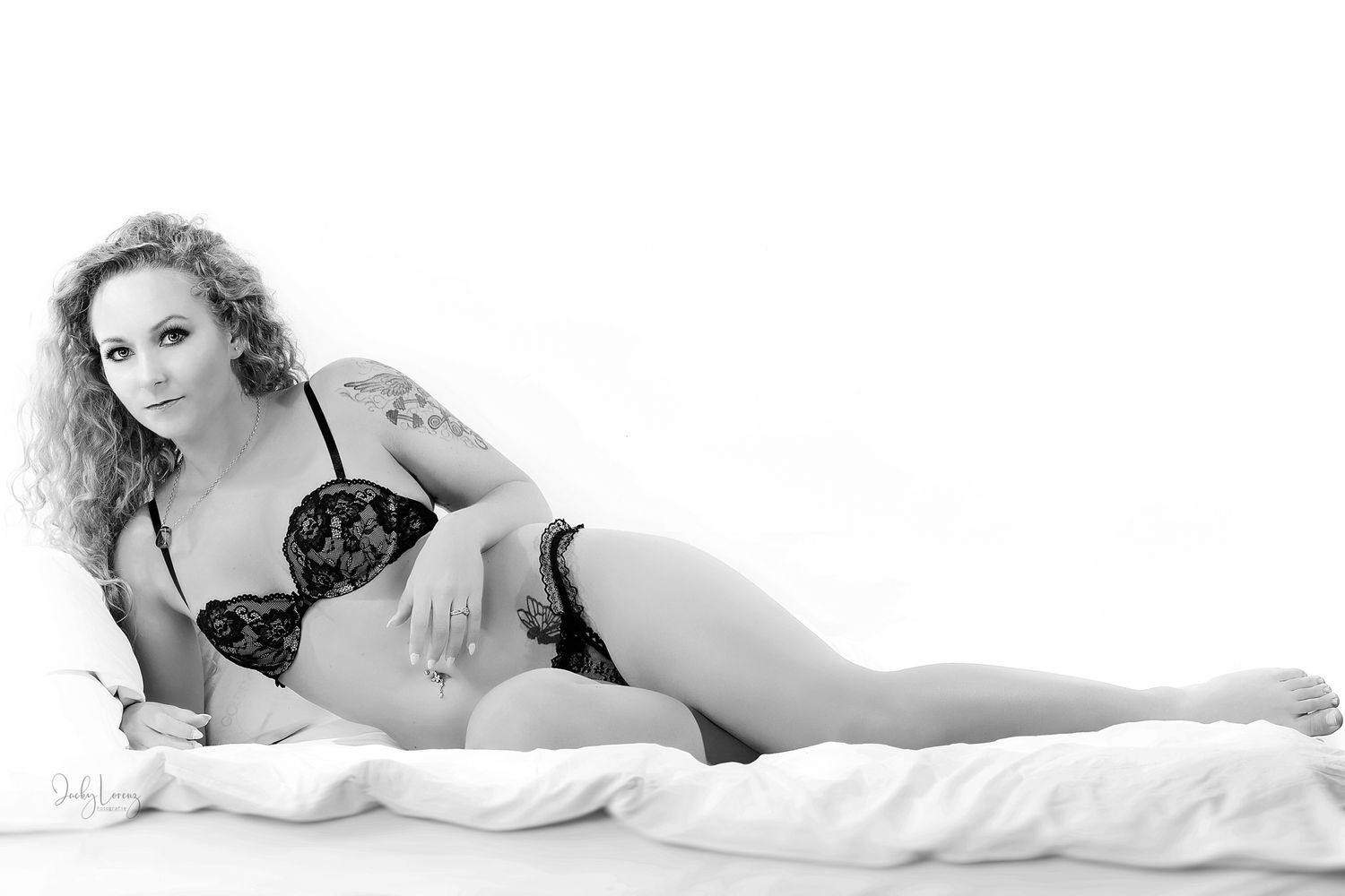 Black and white boudoir photo of someone reclining on white bedding in a romantic pose.