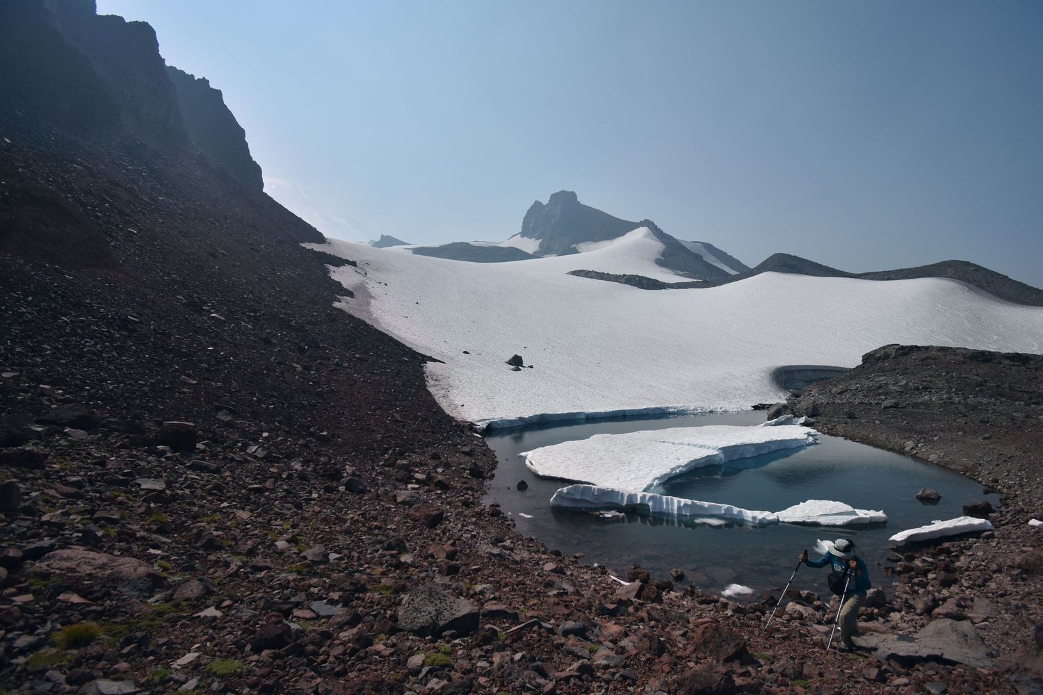 An ice-locked lake on Mount Rainier sits with large floating blocks of ice on the edge of a snowfield. A hiker walks up the rocky hill.