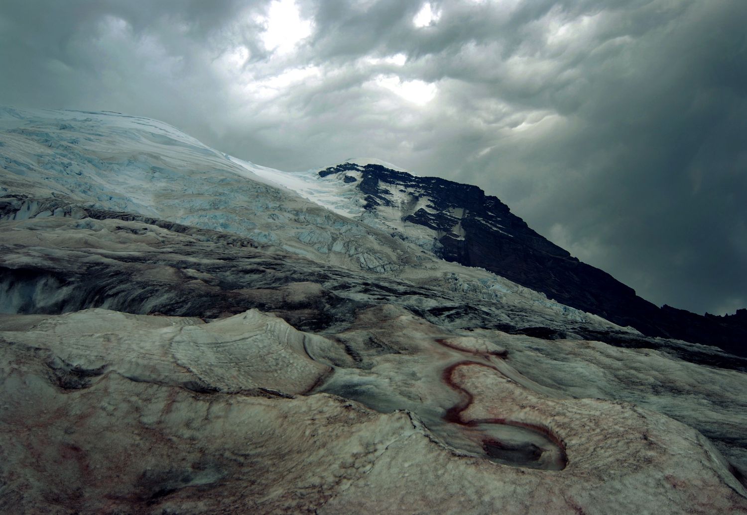 Stormy clouds gather above Mount Rainier in this view from near Camp Schurman that shows watermelon algea on the Winthrop glacier.