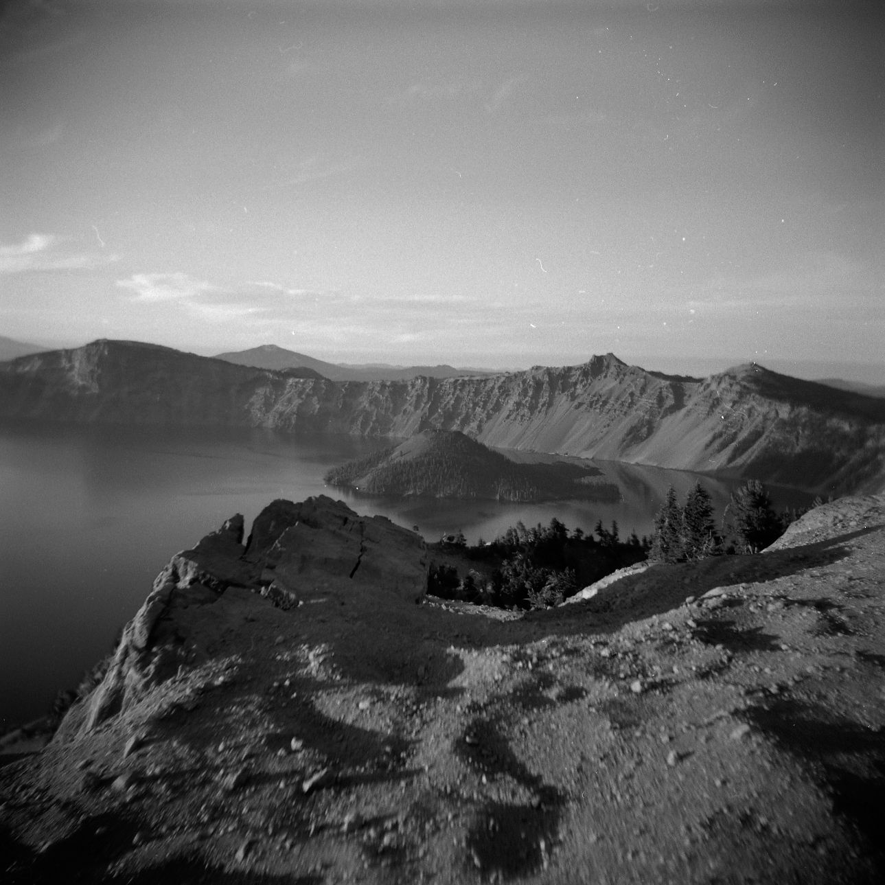 A black and white film Holga photograph of the rim of Crater Lake in Oregon, with Wizard Island visible near the center.