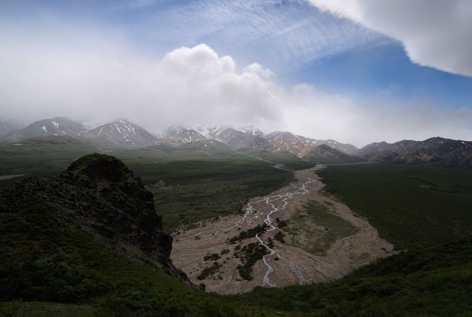 An epic view of braided gravel rivers cutting across open mossy tundra with interestingly foggy partial clouds from which mountains emerge.