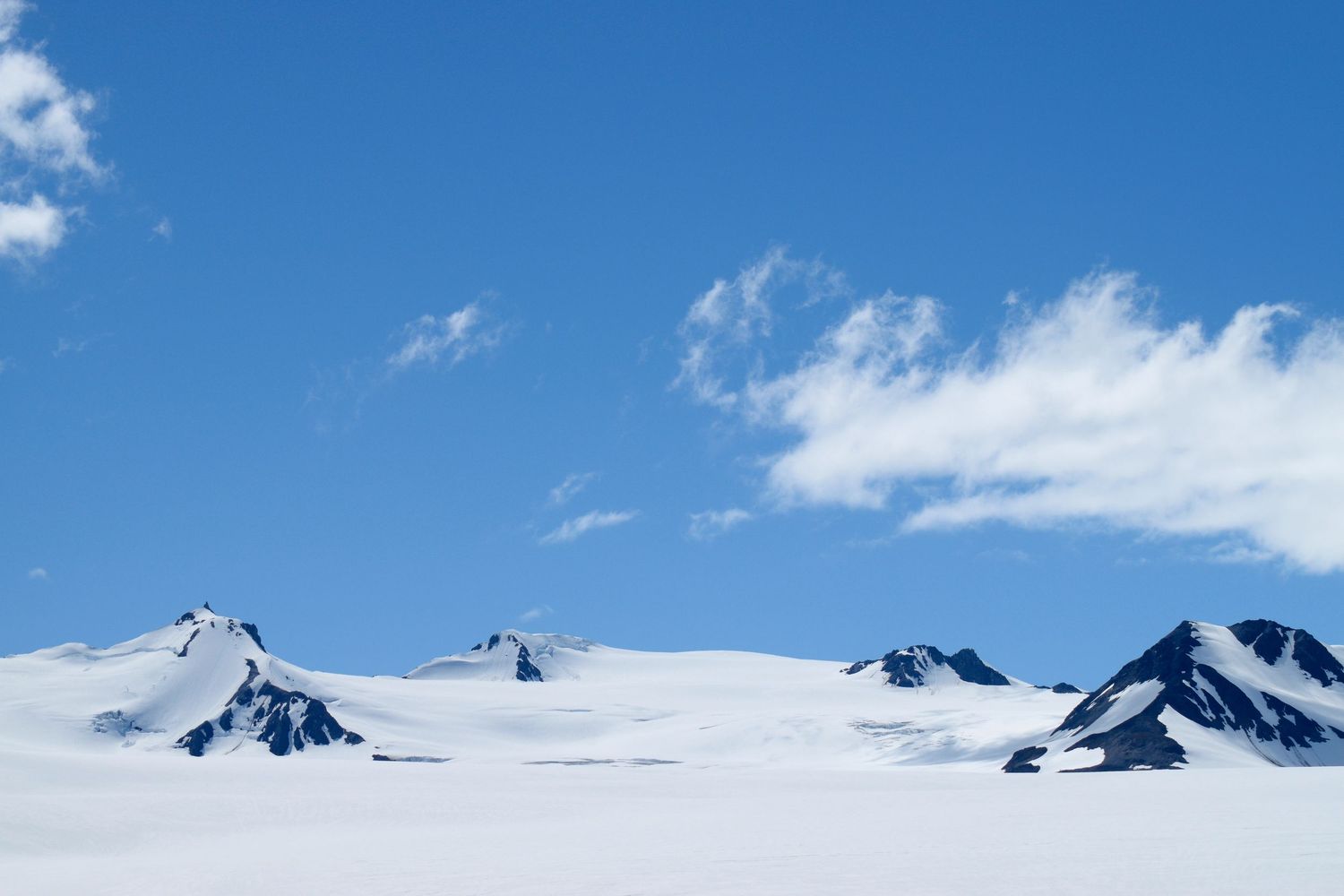 A serene sky with pure a white ice sheet ice and nunataks, or little tops of mountains poking out the ice beneath the few clouds in the sky.