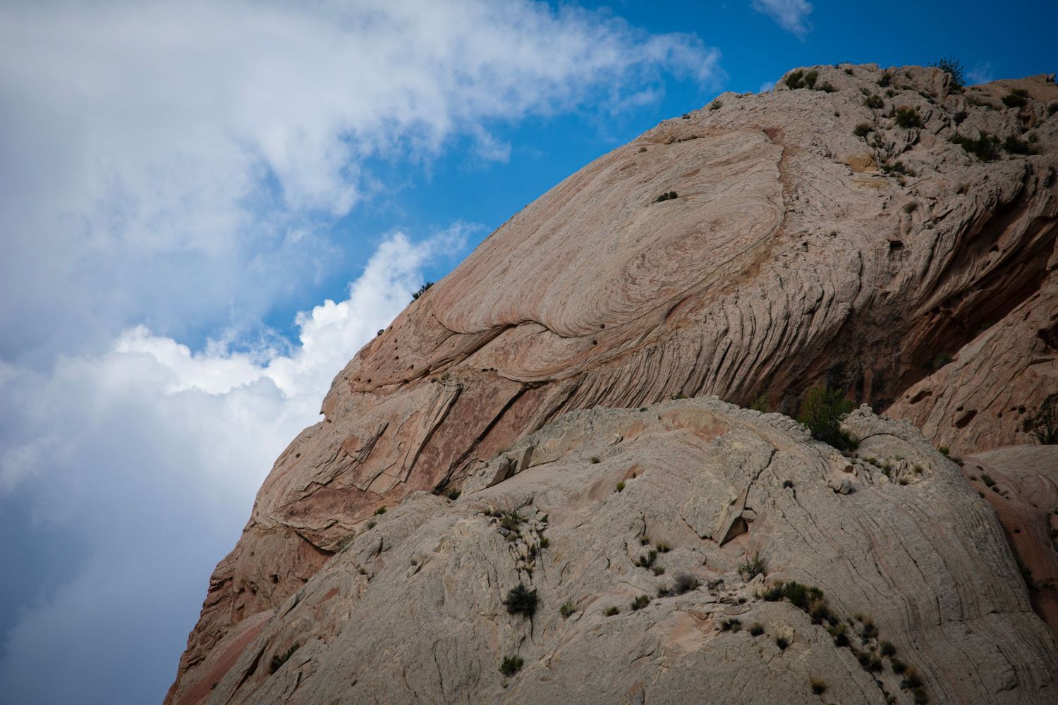 A curved almost egg-like formation of tilted sandstone that is the edge of a huge cliff curves across the frame in front of sky and clouds.