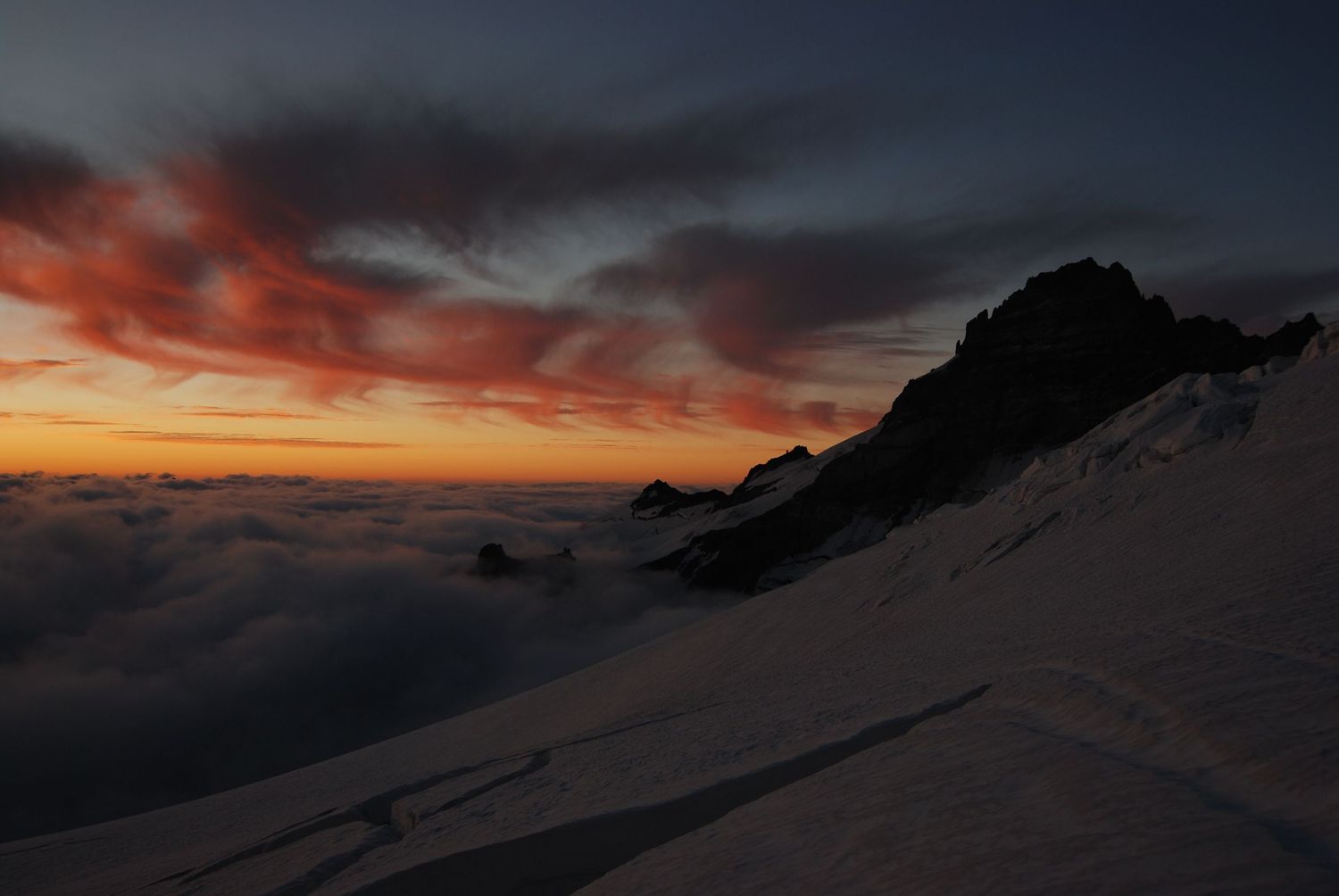 A dramatic red cloud and orange horizon sunset with the dark peak of Little Tahoma and the Emmons glacier below on Mount Rainier.
